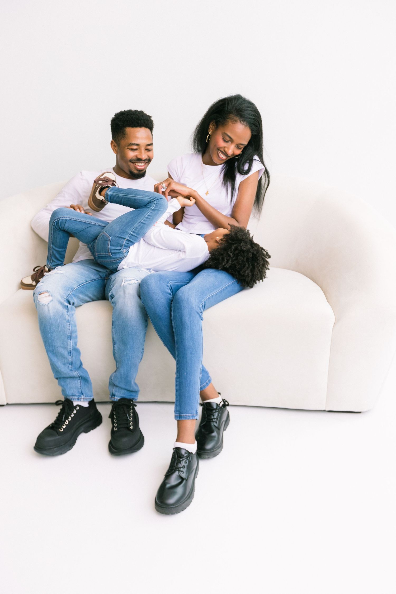 Happy Black family of three sitting on a white couch, wearing matching white shirts and jeans, laughing together.