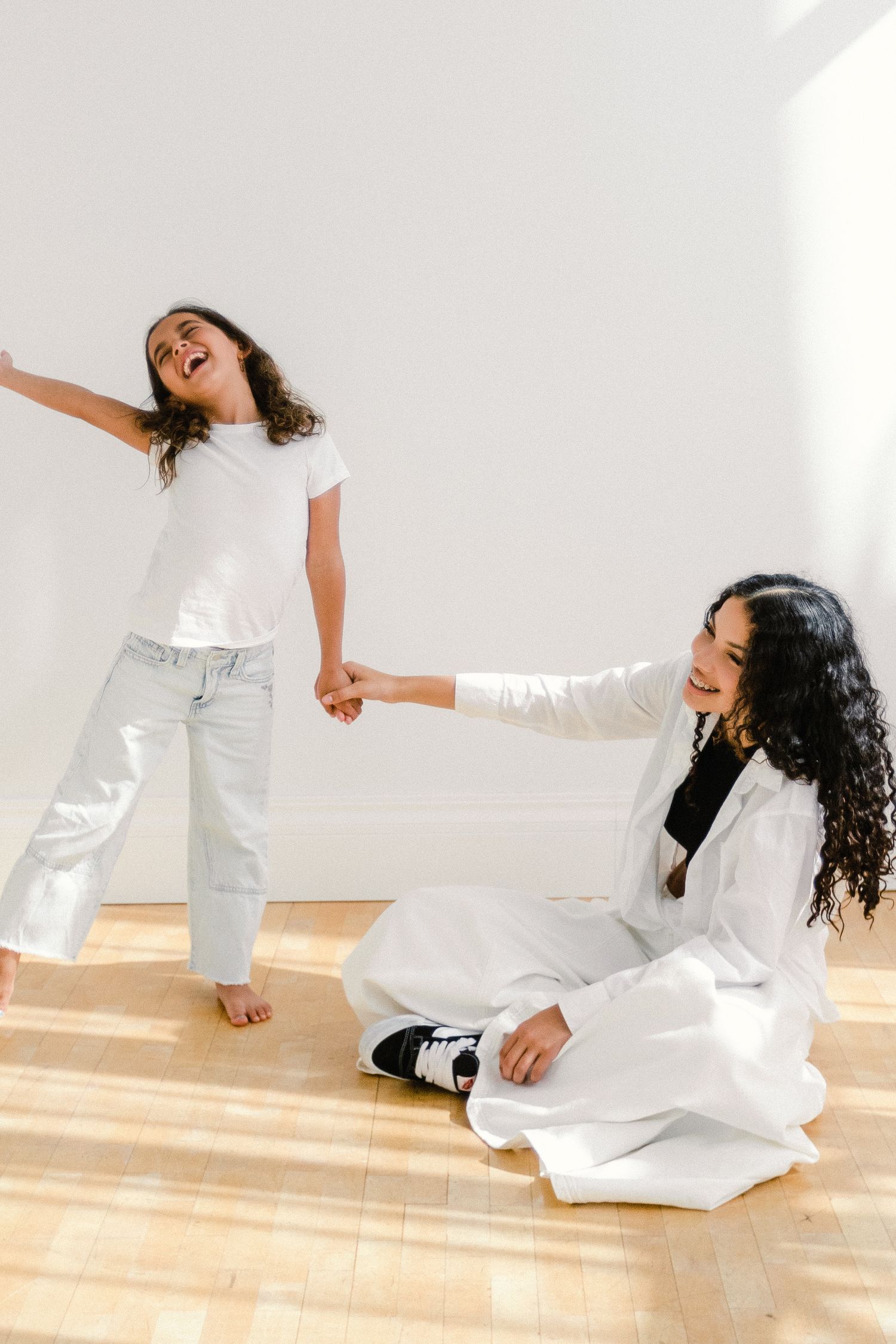 A woman sitting on a hardwood floor holds hands with a laughing young girl in a bright white room.