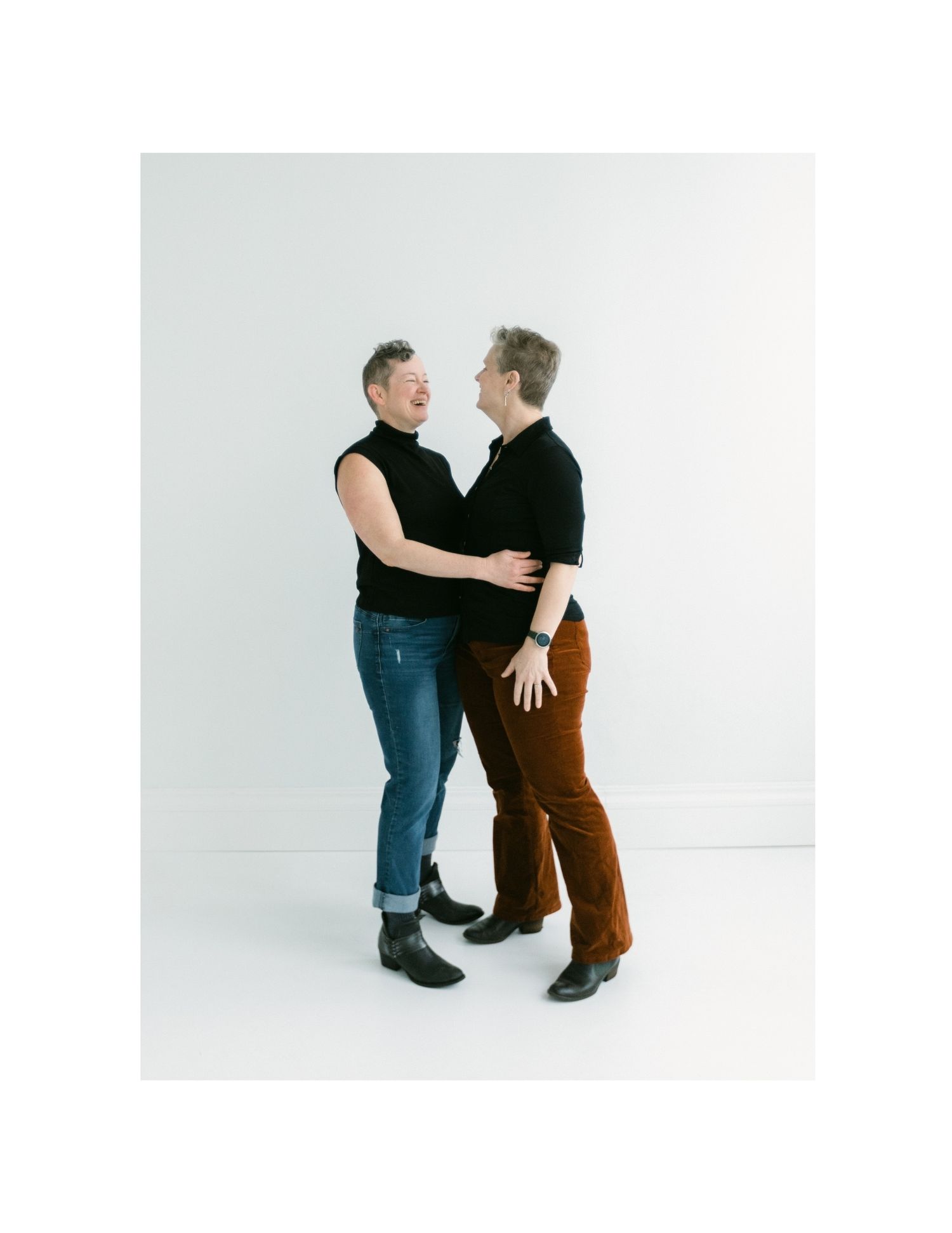 Two women in black tops smile and embrace each other lovingly in a bright white studio.