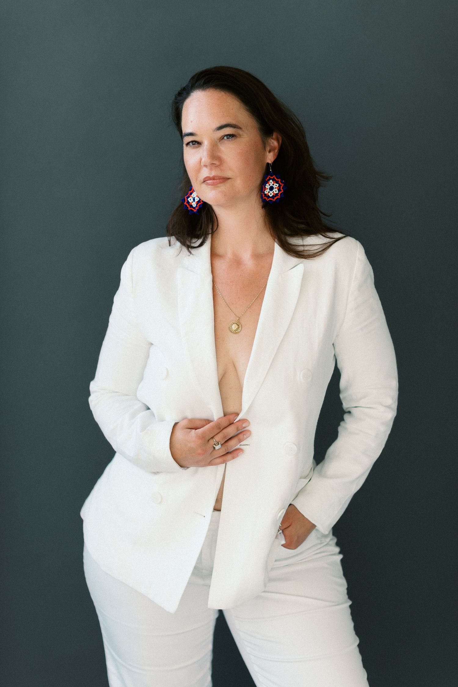 Woman in white blazer suit with pink statement earrings and gold necklace posing against dark gray background.