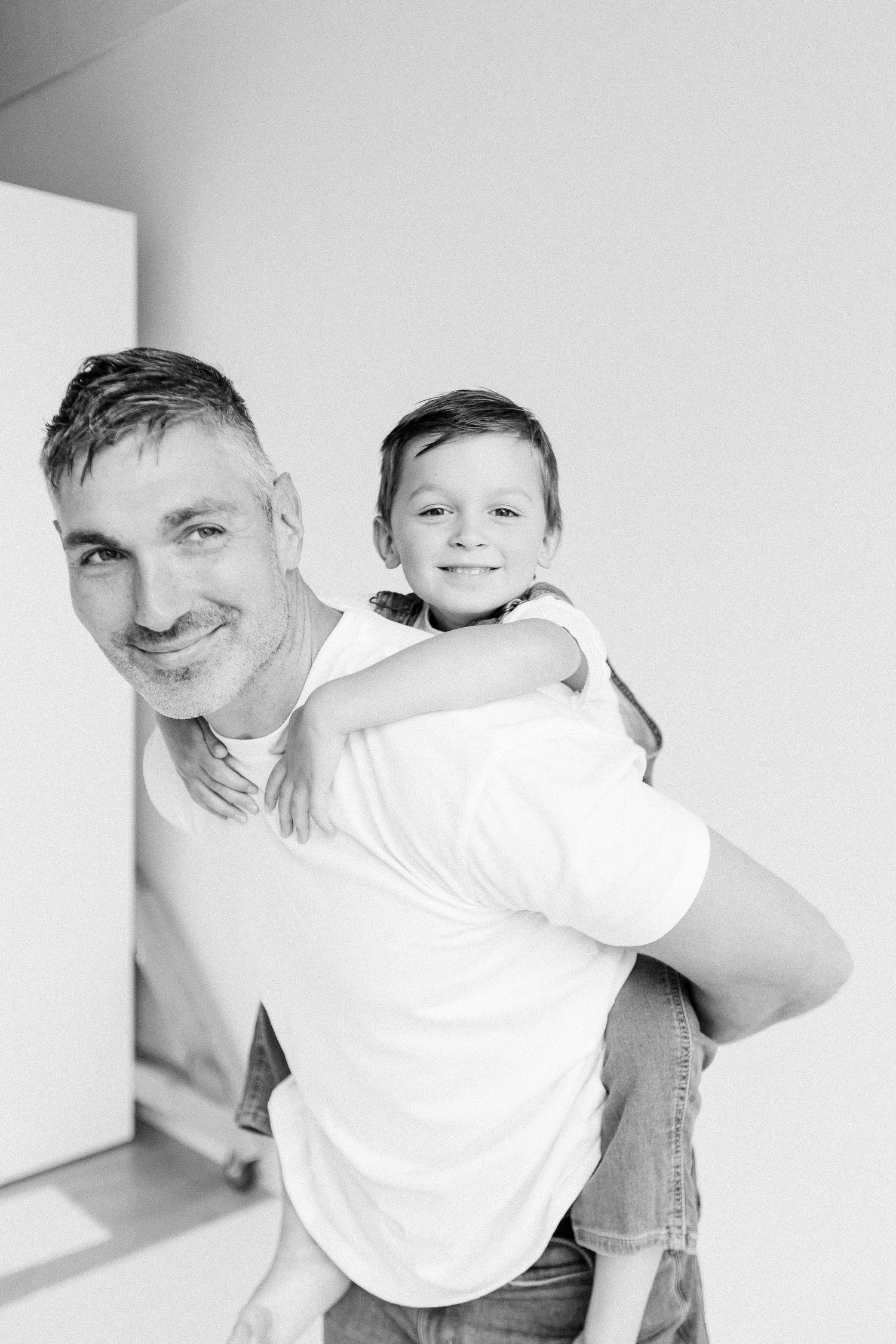 Black and white photo of a smiling man giving a happy young boy a piggyback ride indoors.