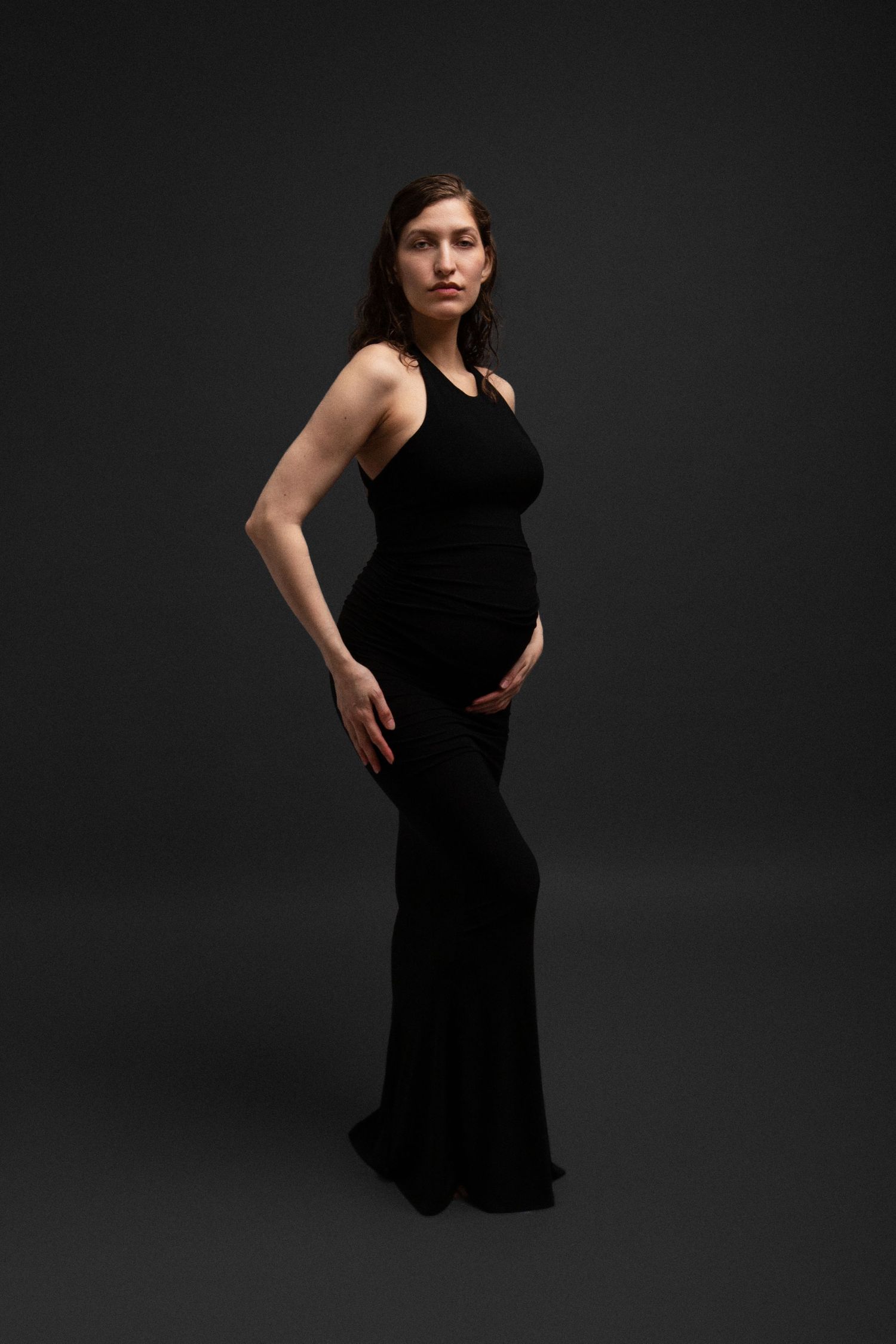 Pregnant woman in elegant black sleeveless maxi dress posing against dark studio background.