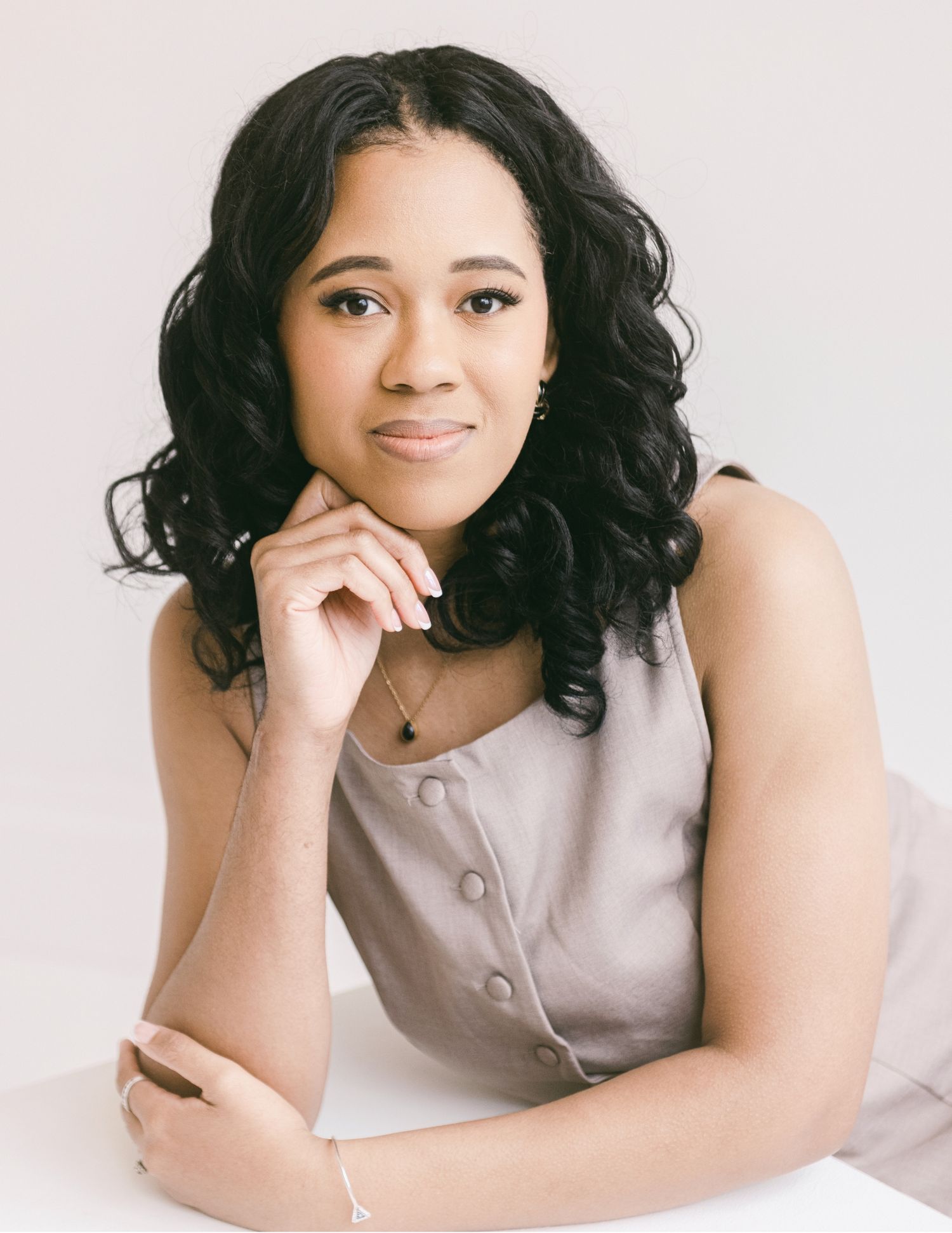 A young woman with curly black hair in a gray sleeveless top poses thoughtfully with her hand near her chin.
