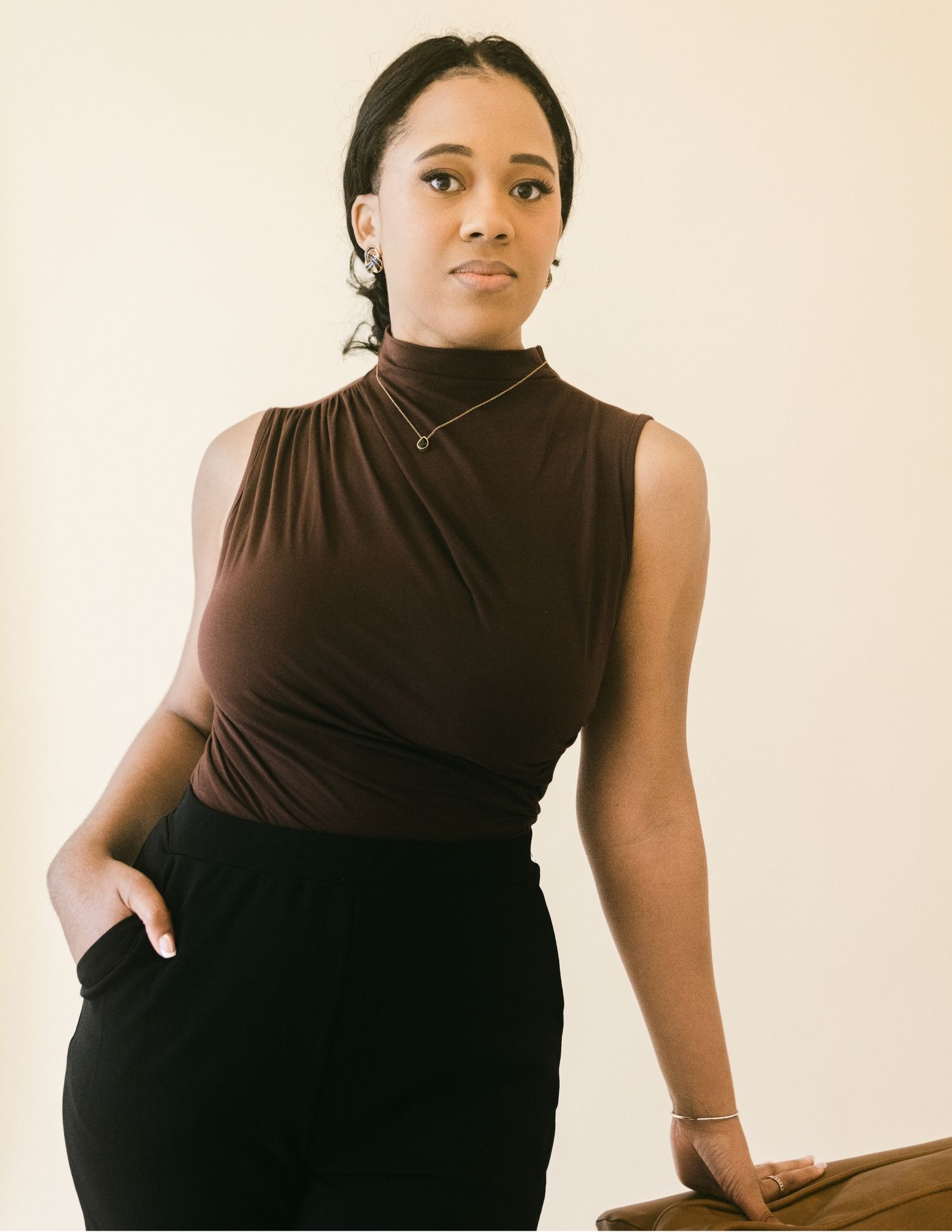 Young woman in a brown sleeveless turtleneck and black pants poses confidently with hand in pocket.