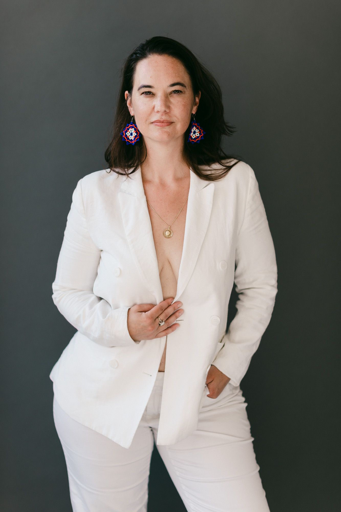 Woman in white blazer and pants with colorful earrings posing confidently against a dark gray background.