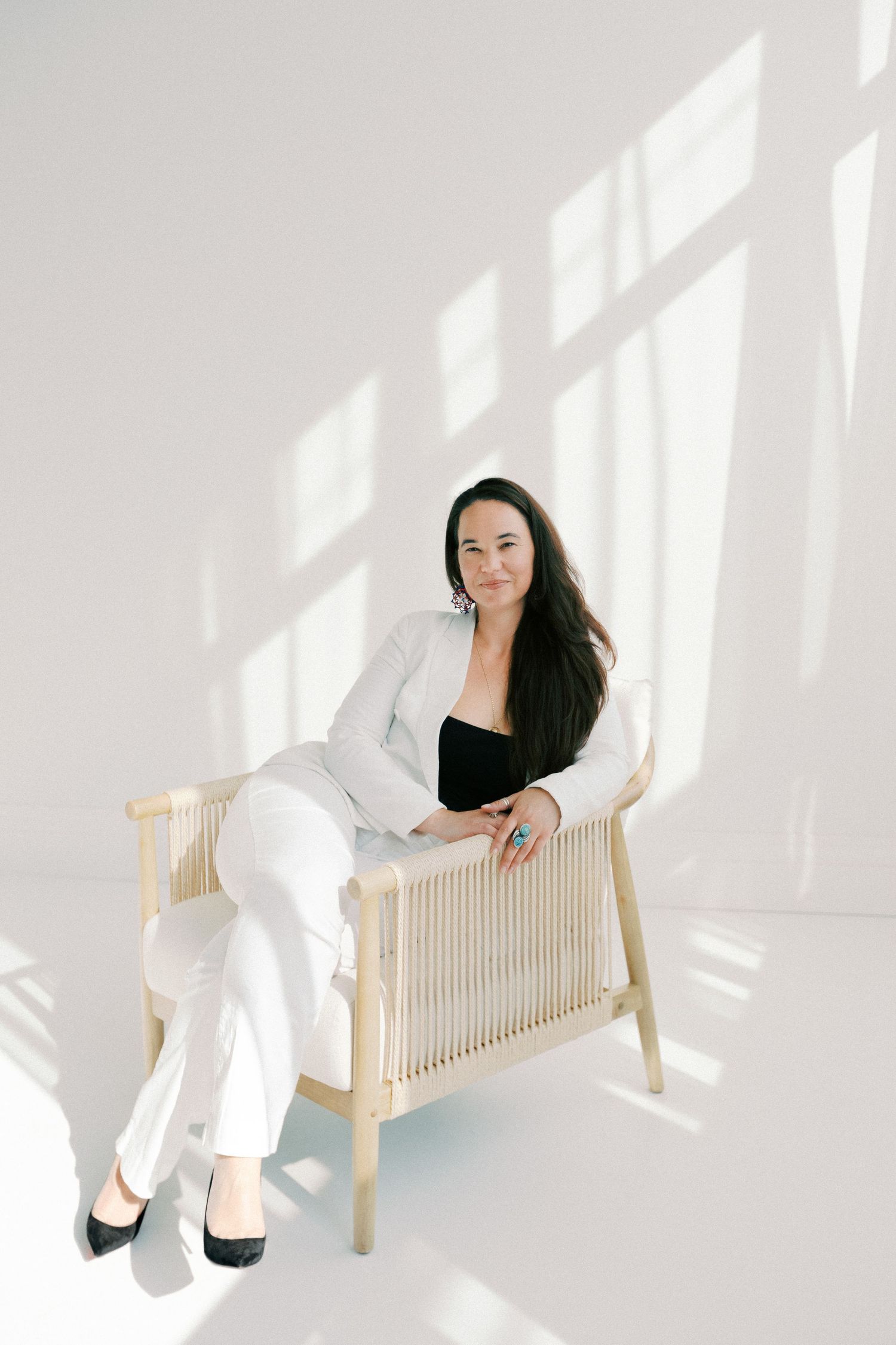 Woman in white suit sitting relaxed on a wooden chair against a bright white wall with natural light shadows.