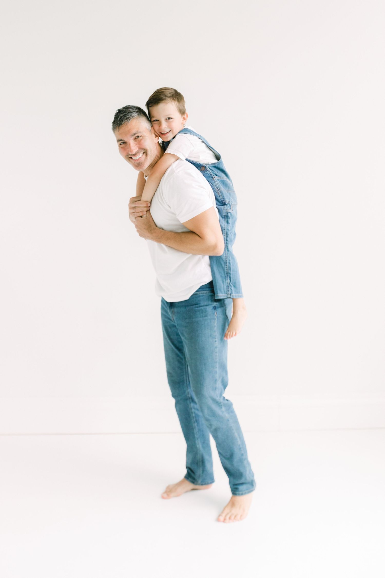 A smiling man gives a happy boy a piggyback ride against a white background, both wearing casual denim outfits.