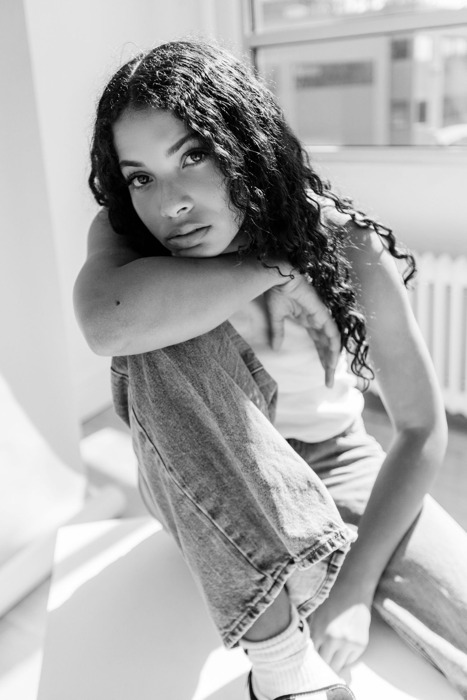 Black and white portrait of a young woman with curly hair sitting casually, wearing jeans and a sleeveless top.