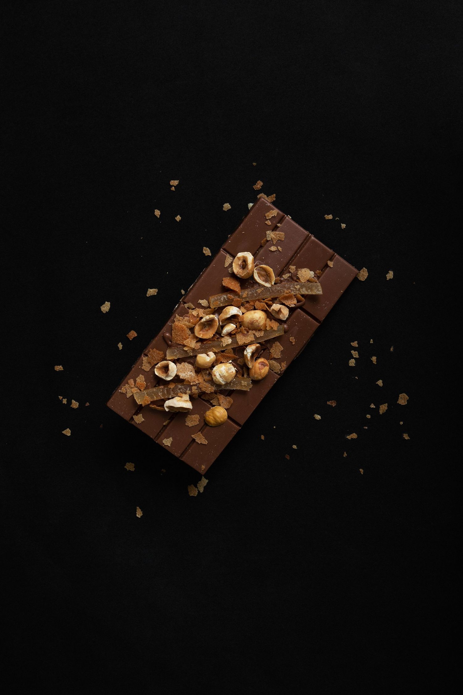 Milk chocolate bar topped with crunchy hazelnuts on a dark black background, viewed from above.