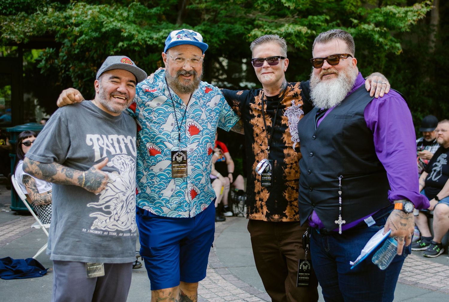 Four tattooed men posing together outdoors at a festival, wearing colorful outfits including Hawaiian shirt and purple jacket.