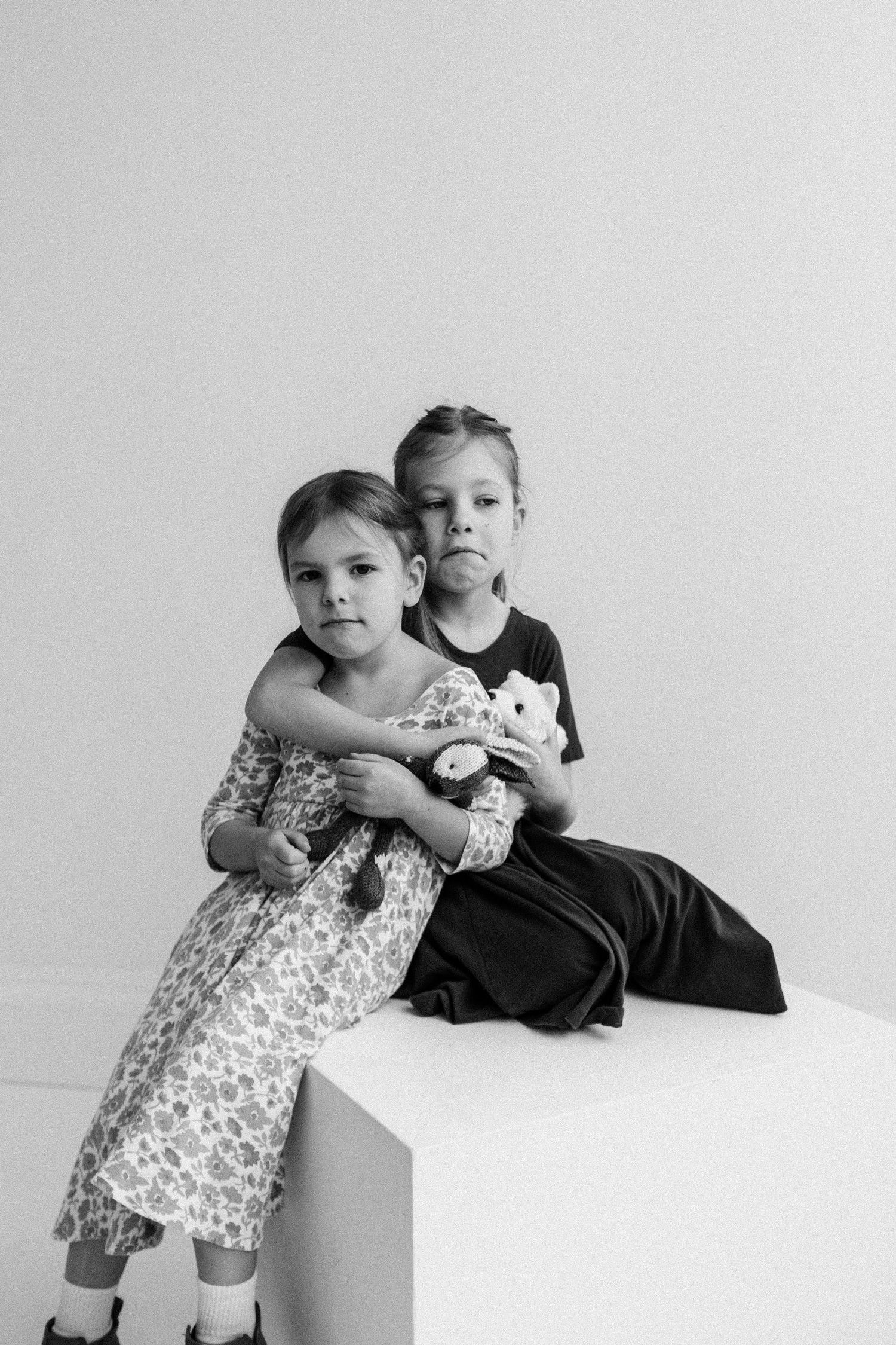 Black and white photo of two young girls hugging while seated on a white cube, holding a stuffed toy.