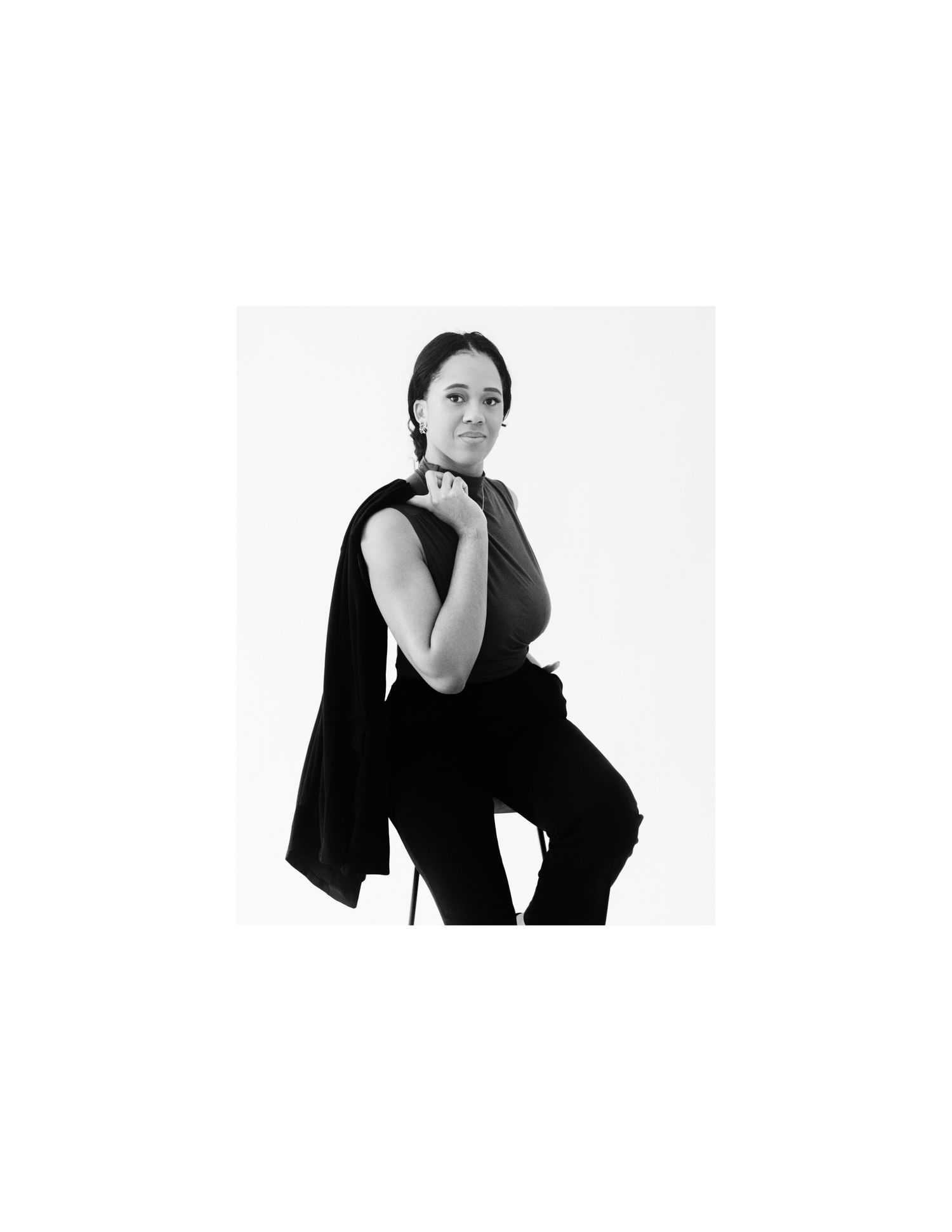 Black and white portrait of a woman in sleeveless top and black pants, posing confidently with jacket draped over shoulder.