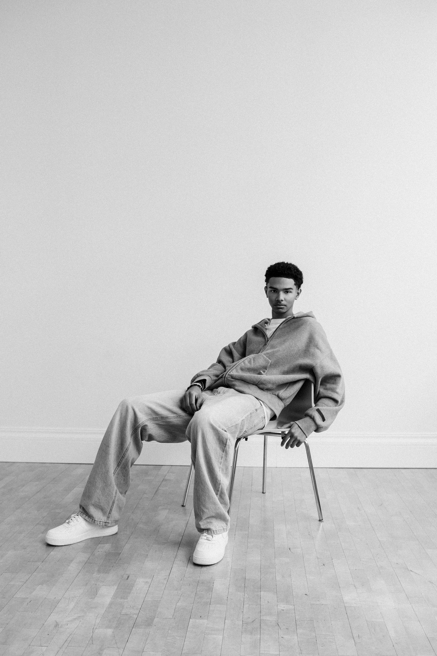 Young man in casual oversized outfit sits on a chair against a white wall in a black and white photo.