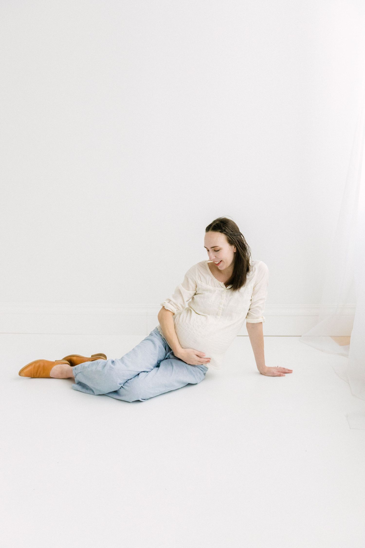 Pregnant woman sitting on white floor, cradling baby bump, wearing cream top, blue jeans, and tan mules.