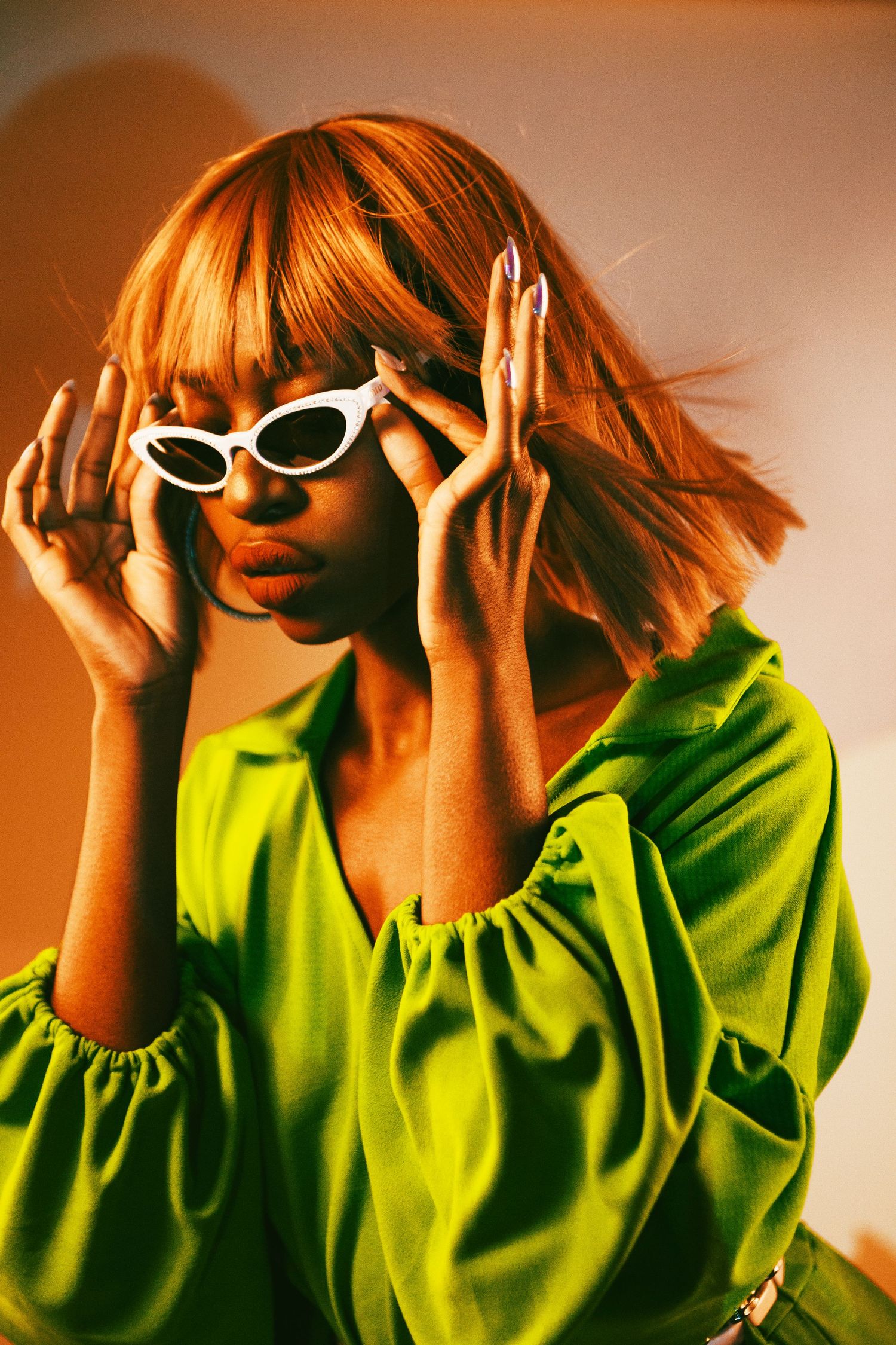 Stylish woman in neon green top with red bob wig adjusting white cat-eye sunglasses under warm studio lighting.