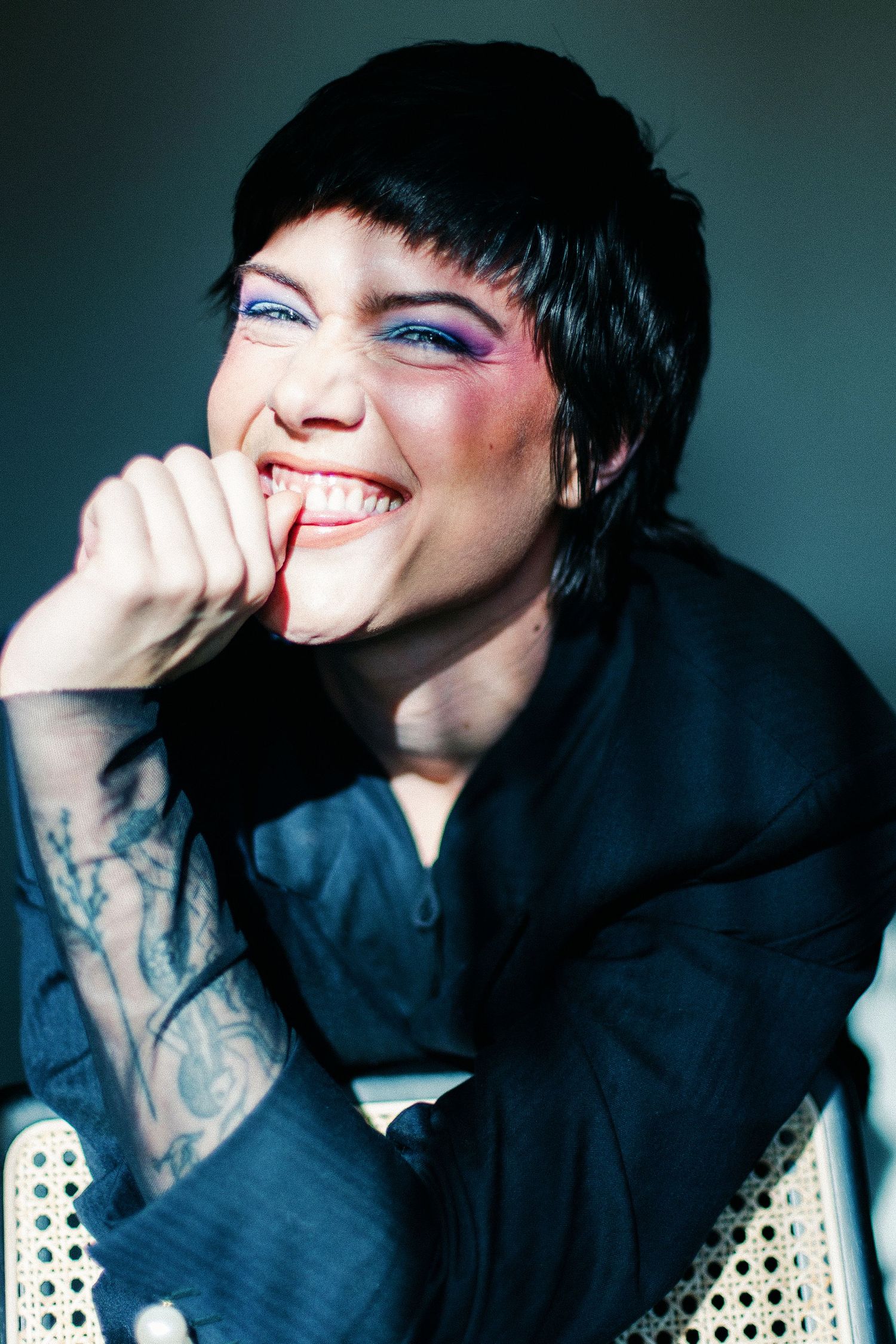 Smiling woman with short black hair, colorful eye makeup, and tattoos wearing a black outfit, resting chin on hand.