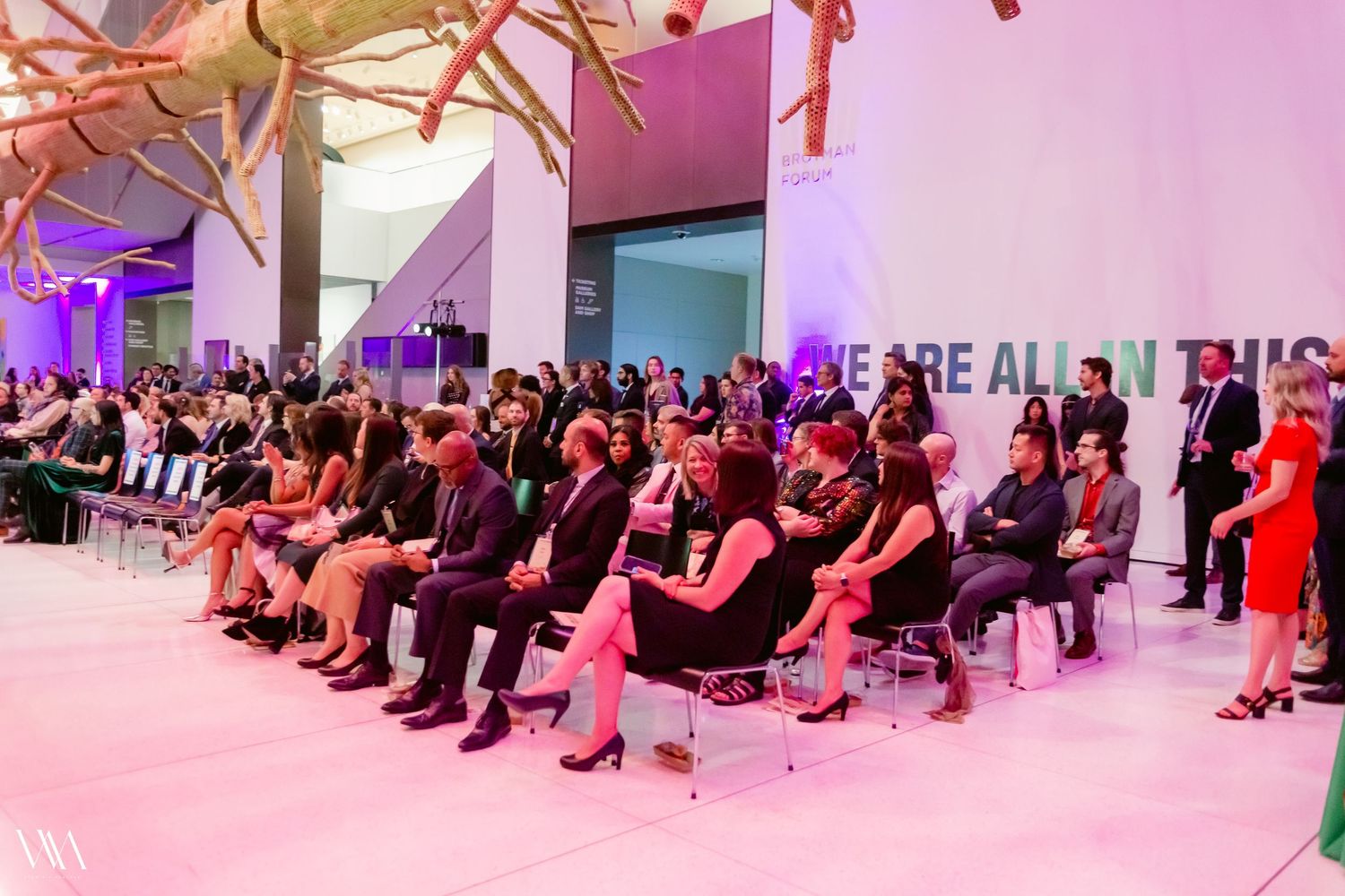 Audience seated at an indoor gala event with purple lighting and a bold motivational message on the wall.