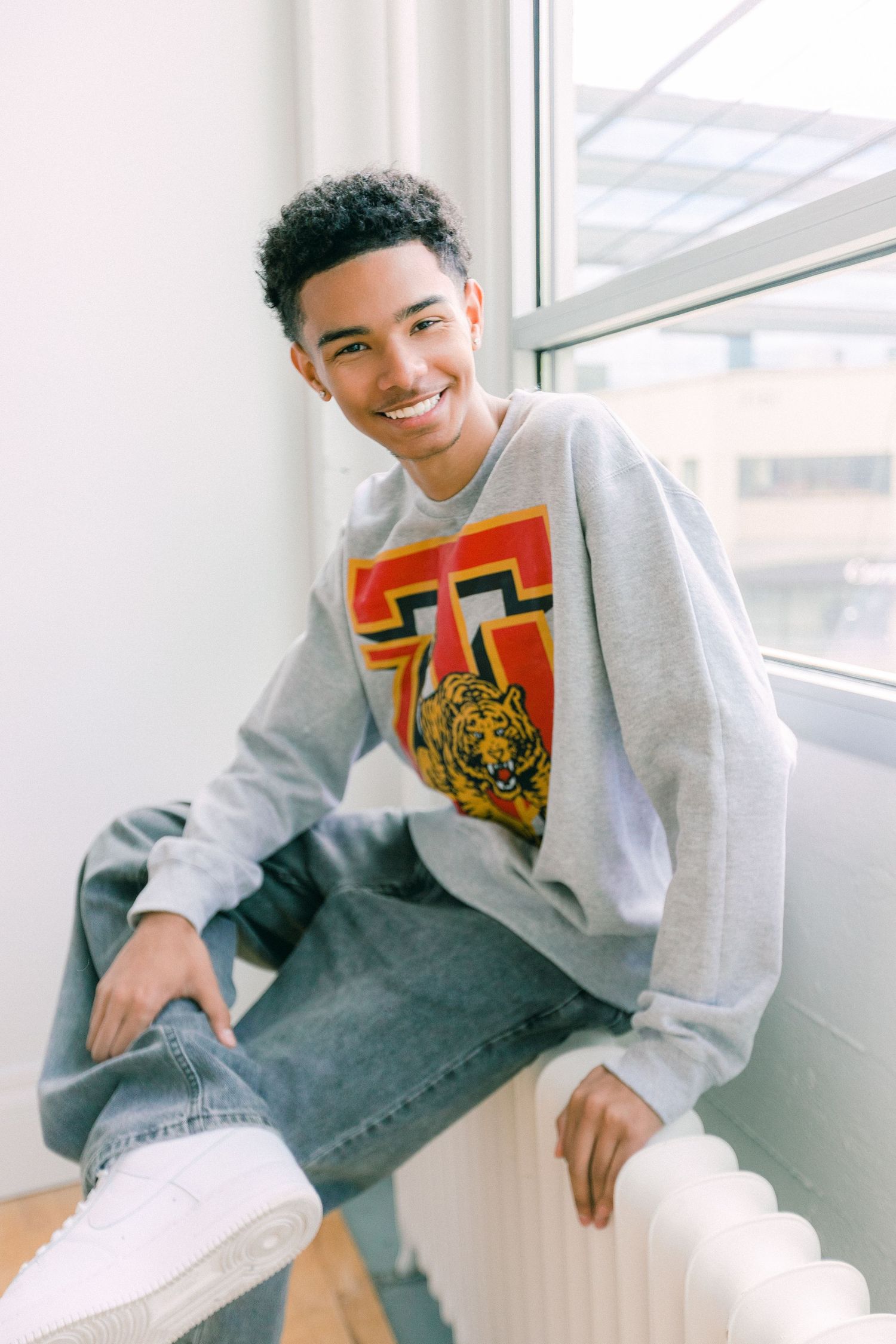 Smiling young man in gray tiger graphic sweatshirt and jeans sits by a bright window on a white radiator.