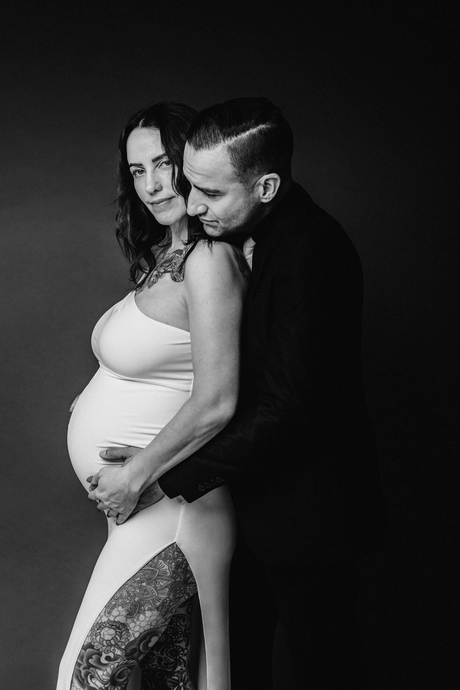 Black and white maternity photo of a pregnant woman in white dress with tattoos, embraced by her partner against dark background.