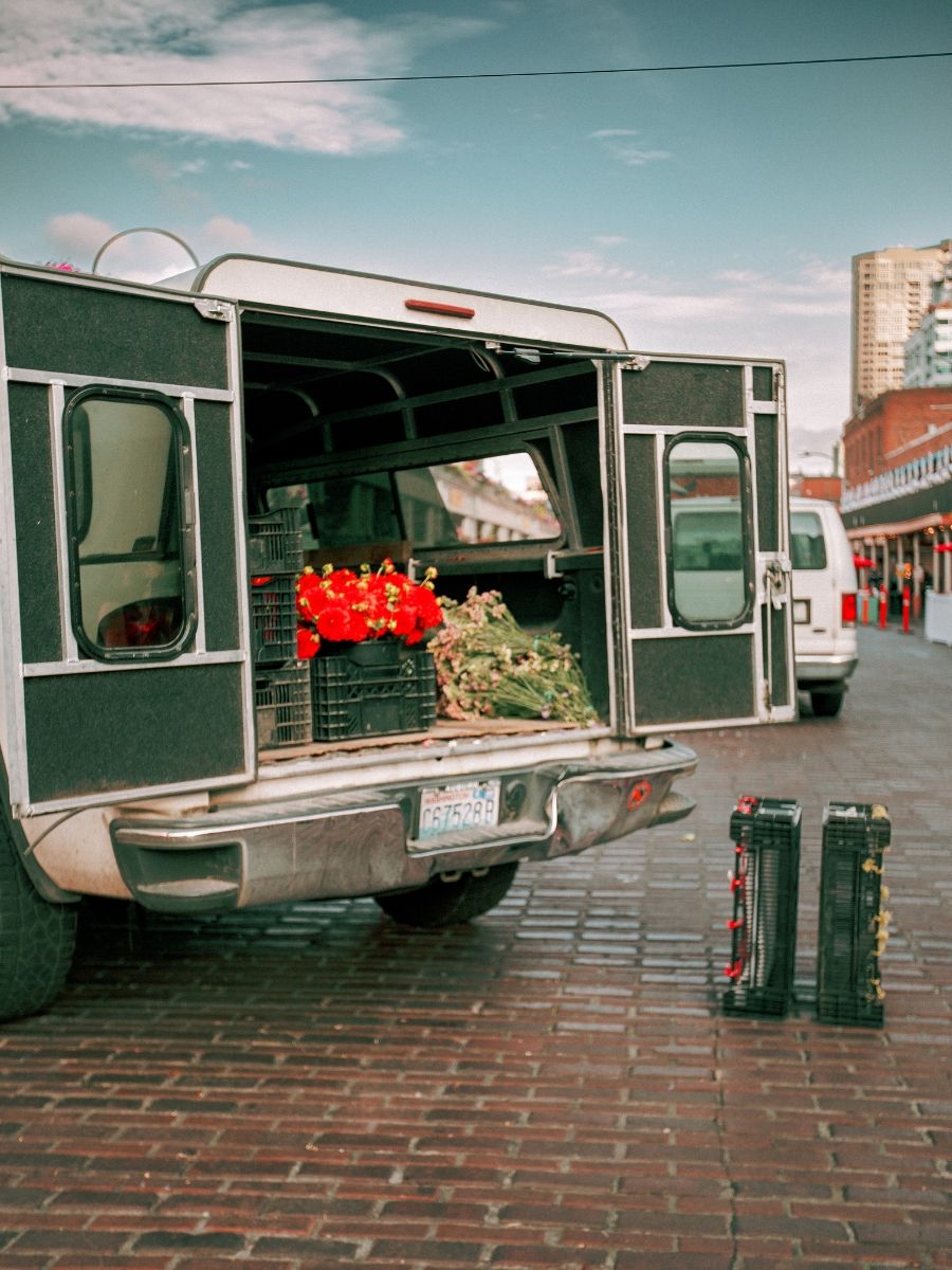 Truck with open rear doors loaded with red flowers and greenery parked on brick street near market.
