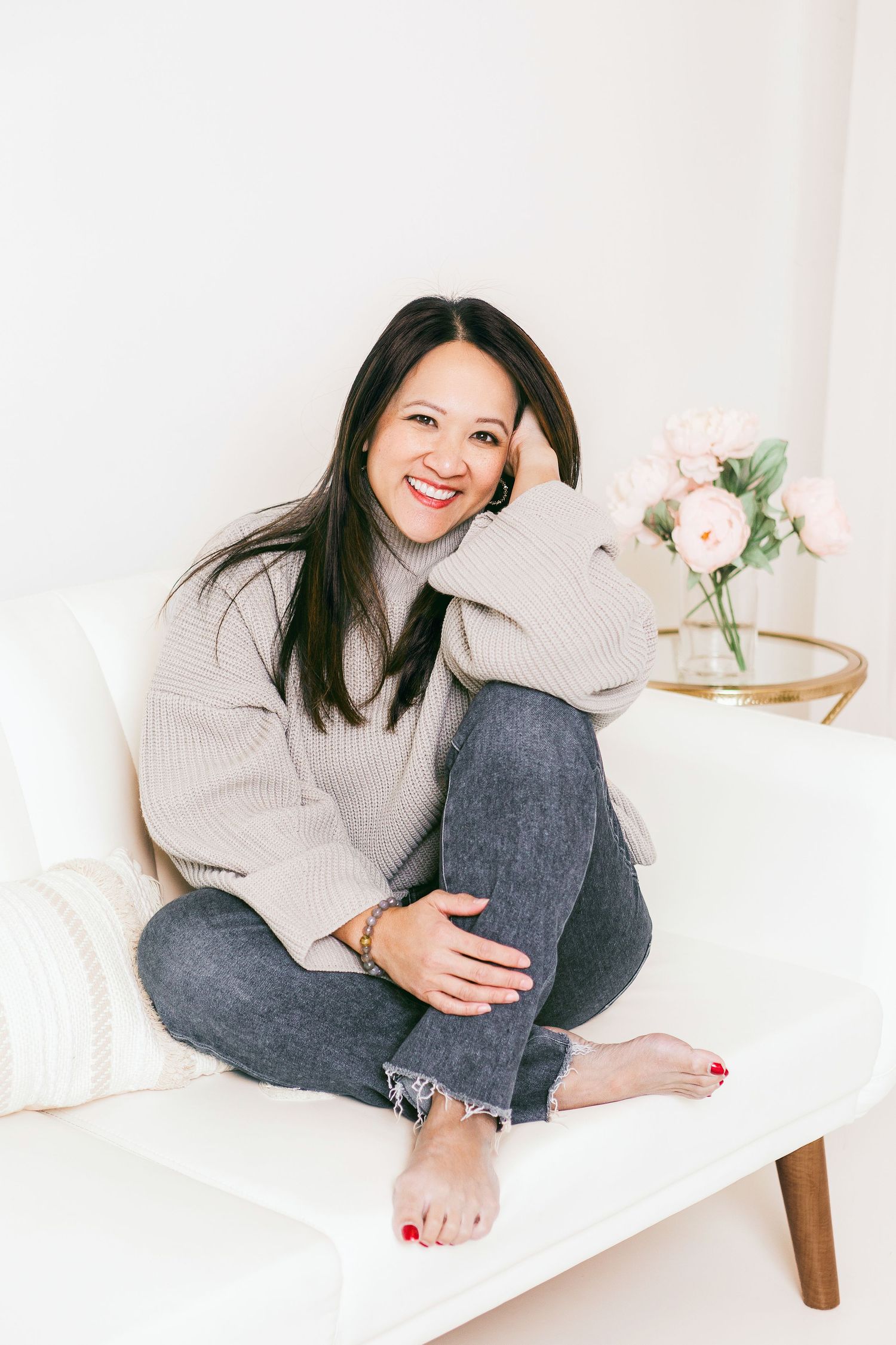 Smiling woman in gray sweater and jeans sitting cross-legged on white sofa with pink flowers in background.