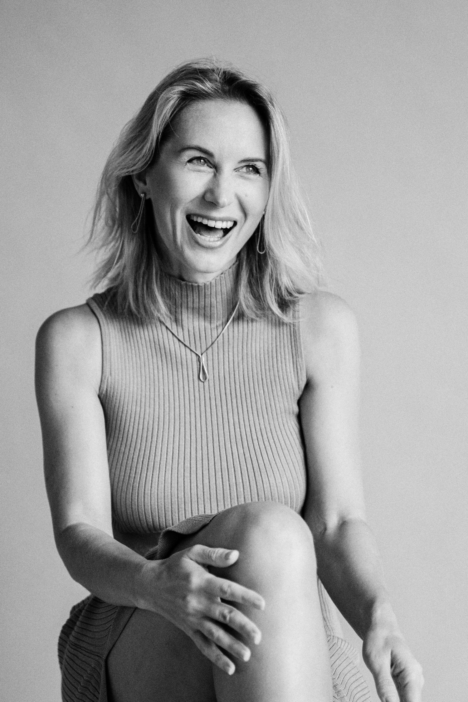 Black and white portrait of a laughing blonde woman in a sleeveless ribbed top, sitting casually with hand on knee.