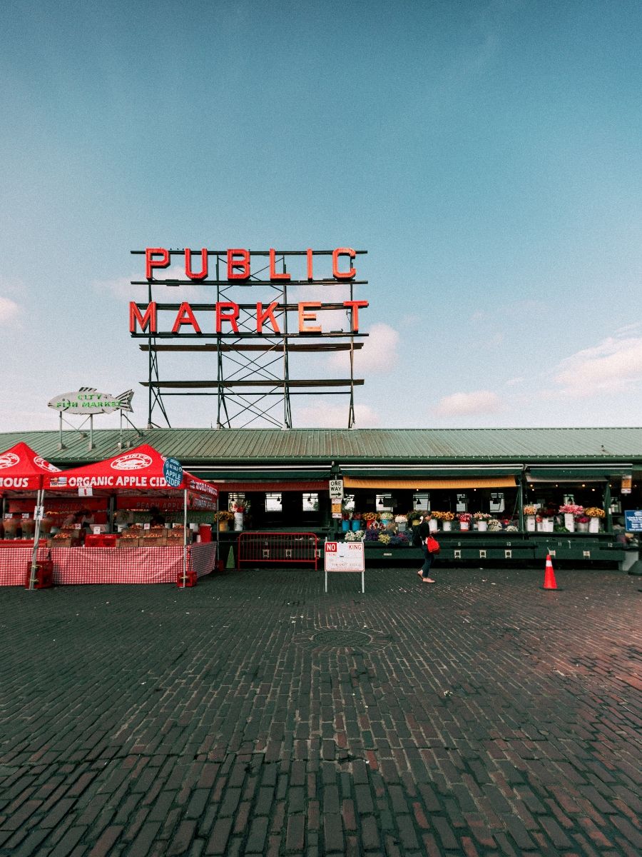 Pike Place Public Market exterior in Seattle, WA with vendors and brick plaza under blue sky.
