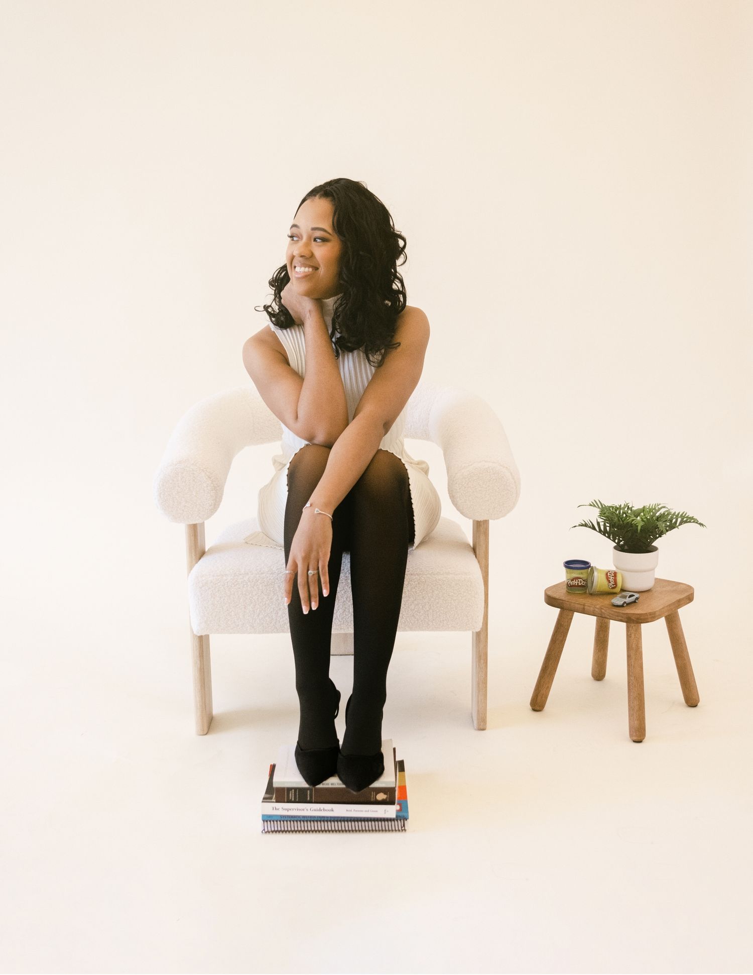 Young woman in gray top and black knee-high socks sits in a white chair beside books and a plant.