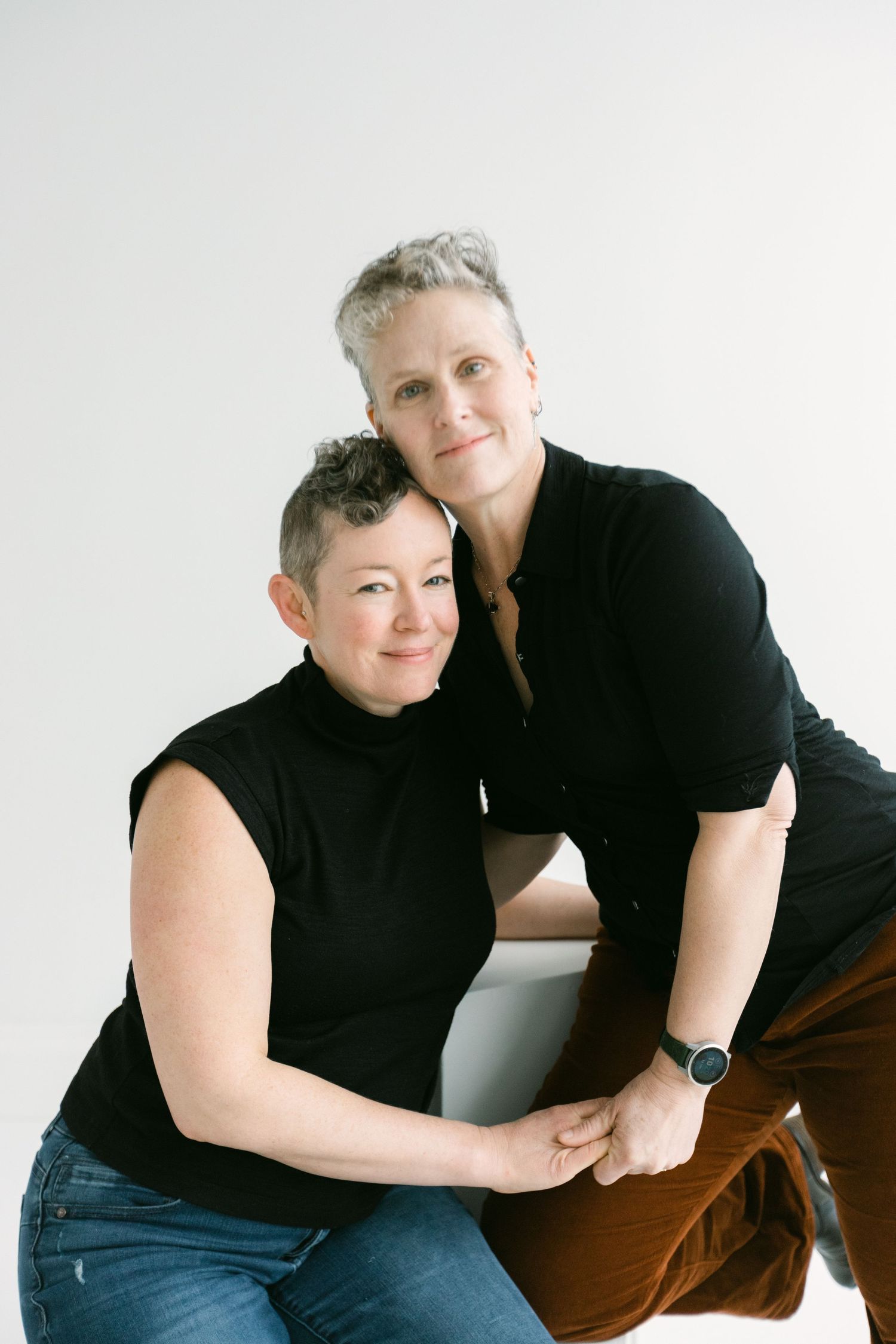 Two women with short gray hair wearing black tops pose together smiling in a bright studio portrait.