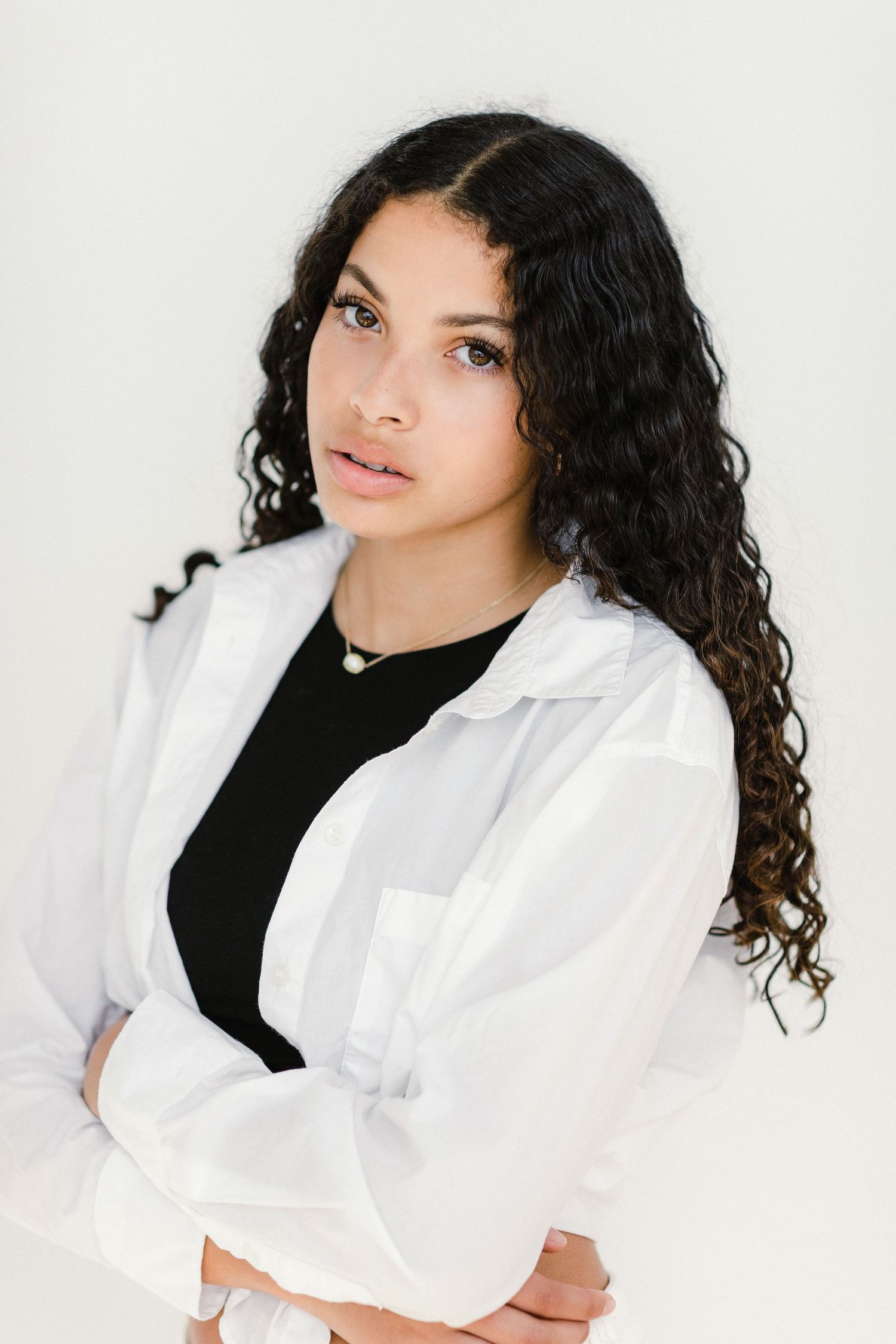 Young woman with curly hair in white shirt and black top posing against light background.