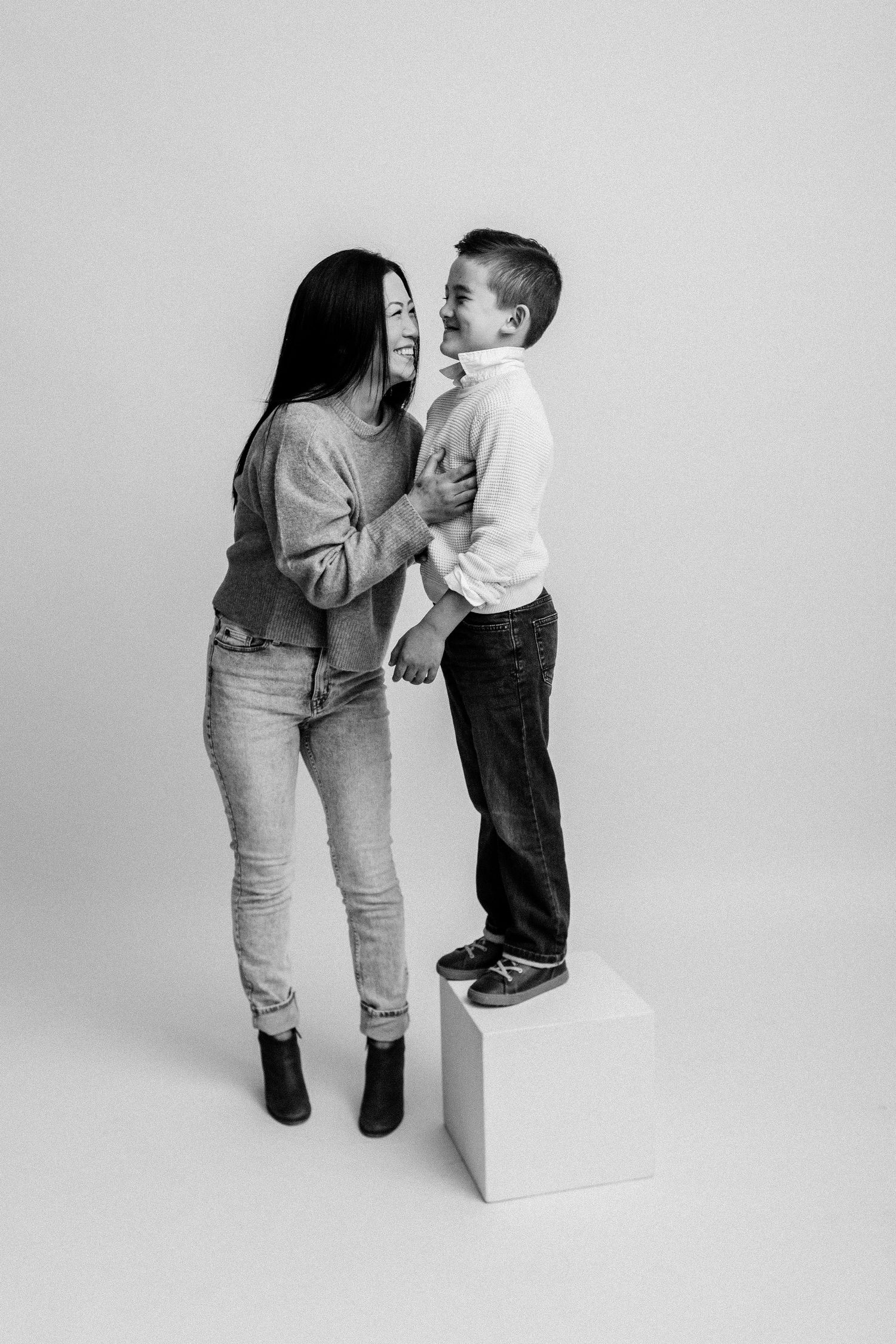 Mom and son share a joyful moment as he stands on a white cube in a studio.