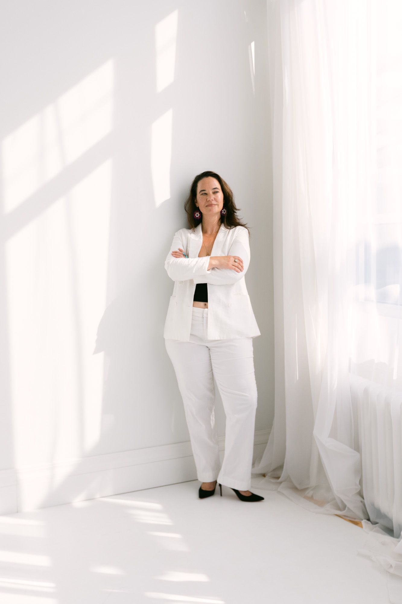 Woman in white blazer and pants stands confidently with arms crossed against a bright white wall.