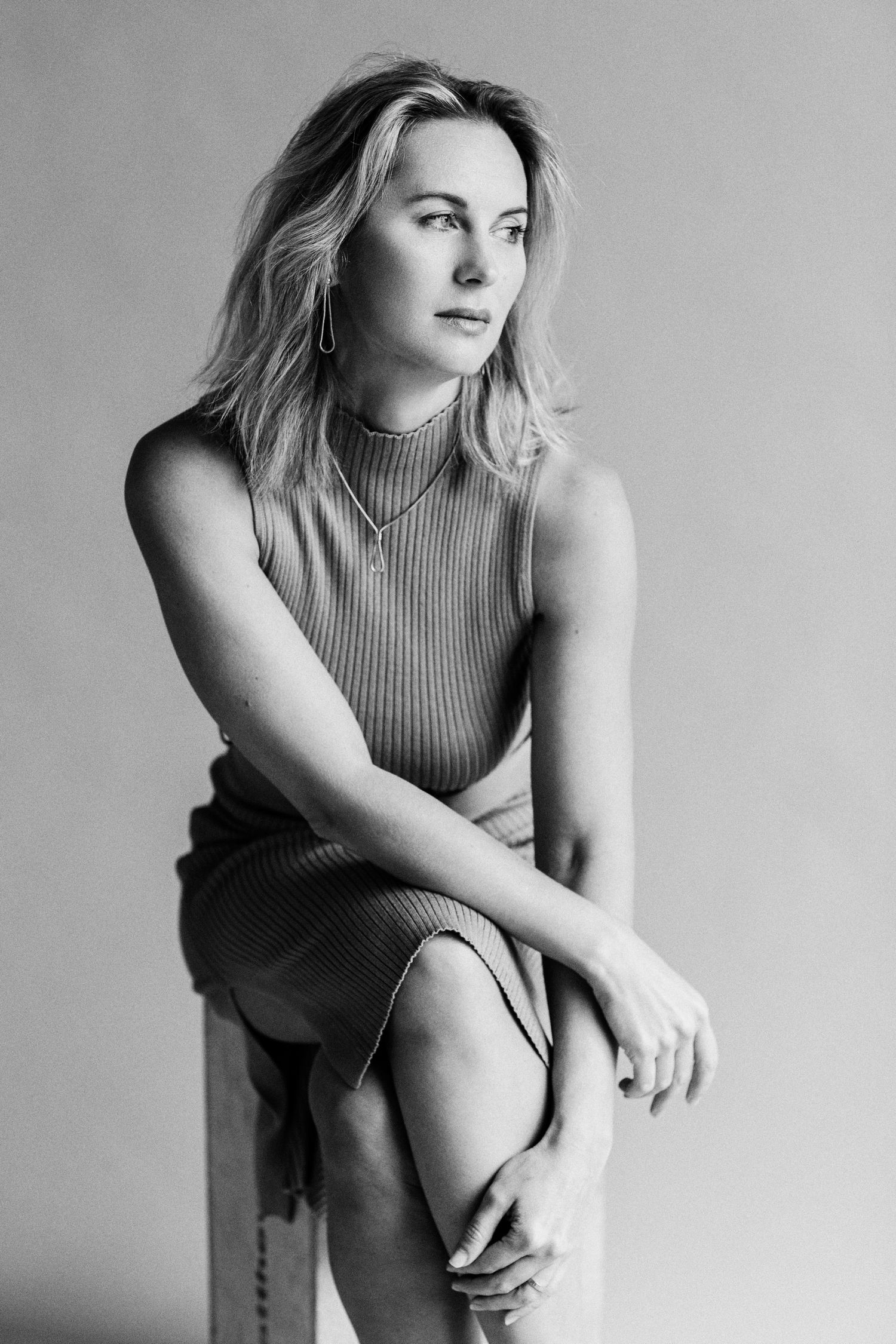 Black and white portrait of a blonde woman in a sleeveless ribbed dress, seated and gazing thoughtfully to the side.