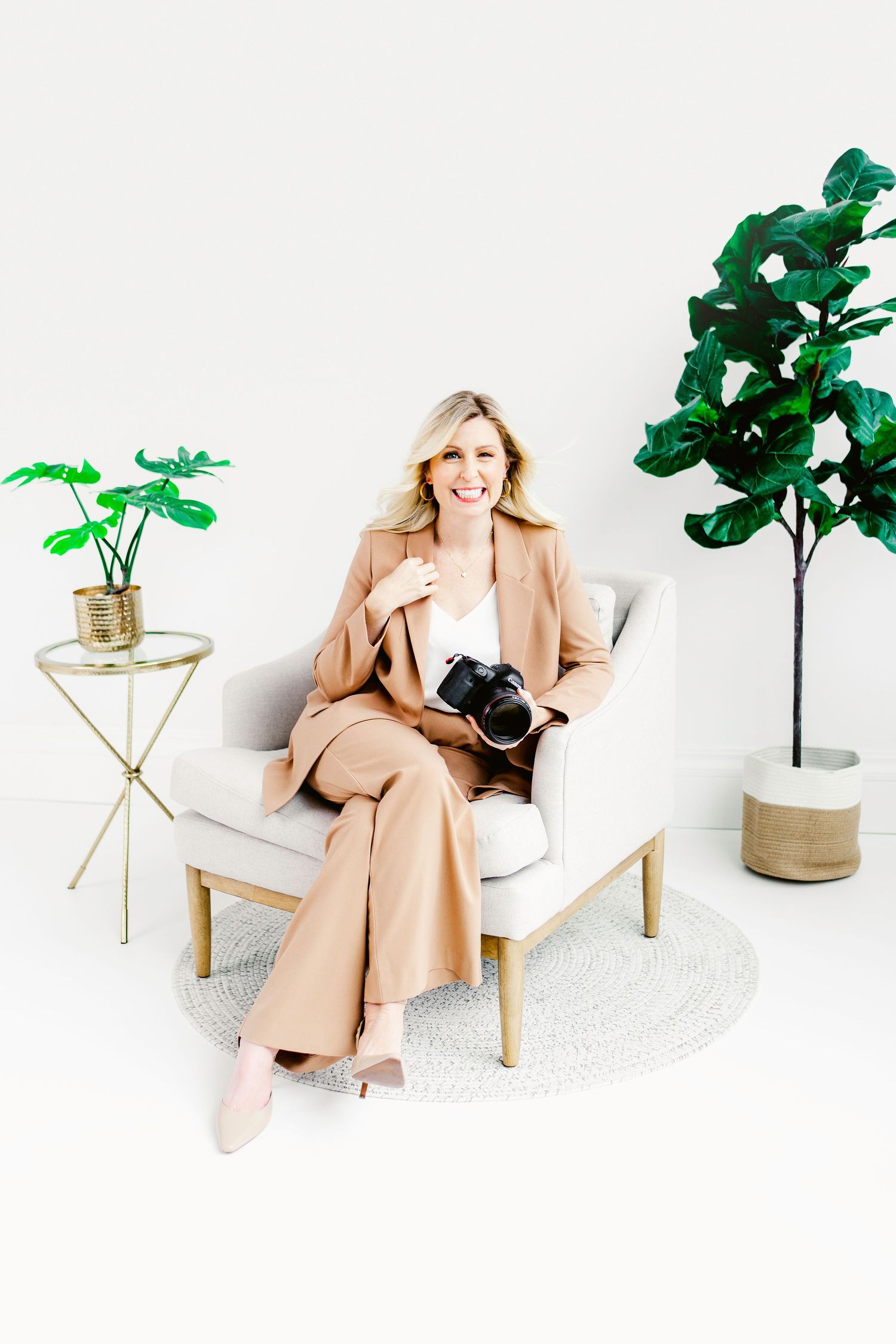Smiling blonde woman in tan suit sitting in white armchair holding camera, surrounded by green plants on white background.