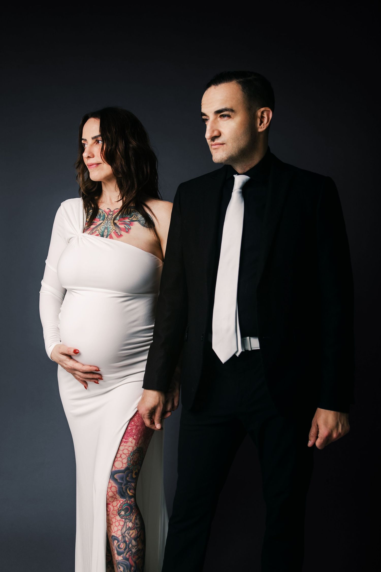 Pregnant woman in white gown with tattoos stands beside man in black suit against dark background.