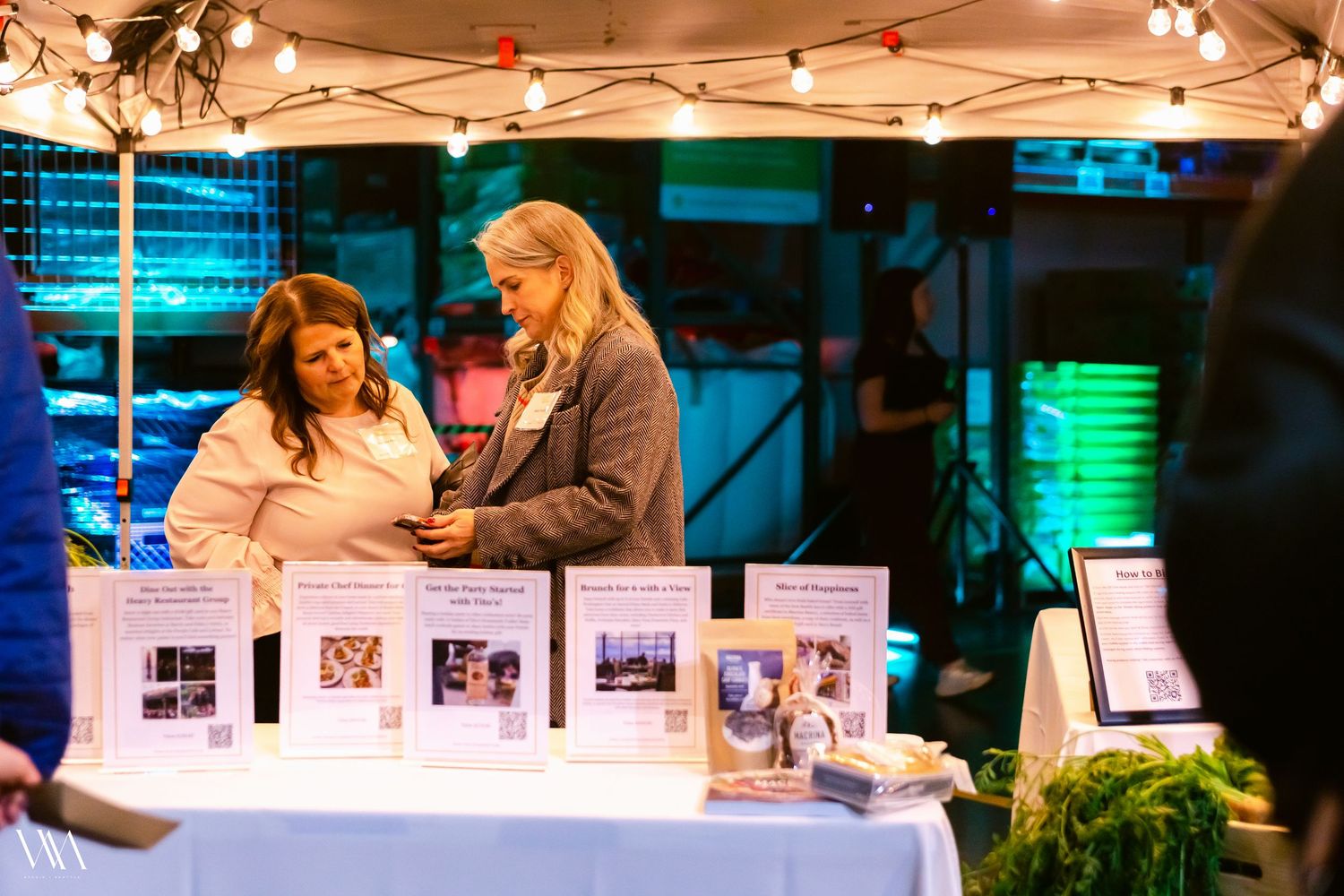 Two women browse informational displays at an outdoor evening vendor booth with string lights overhead.
