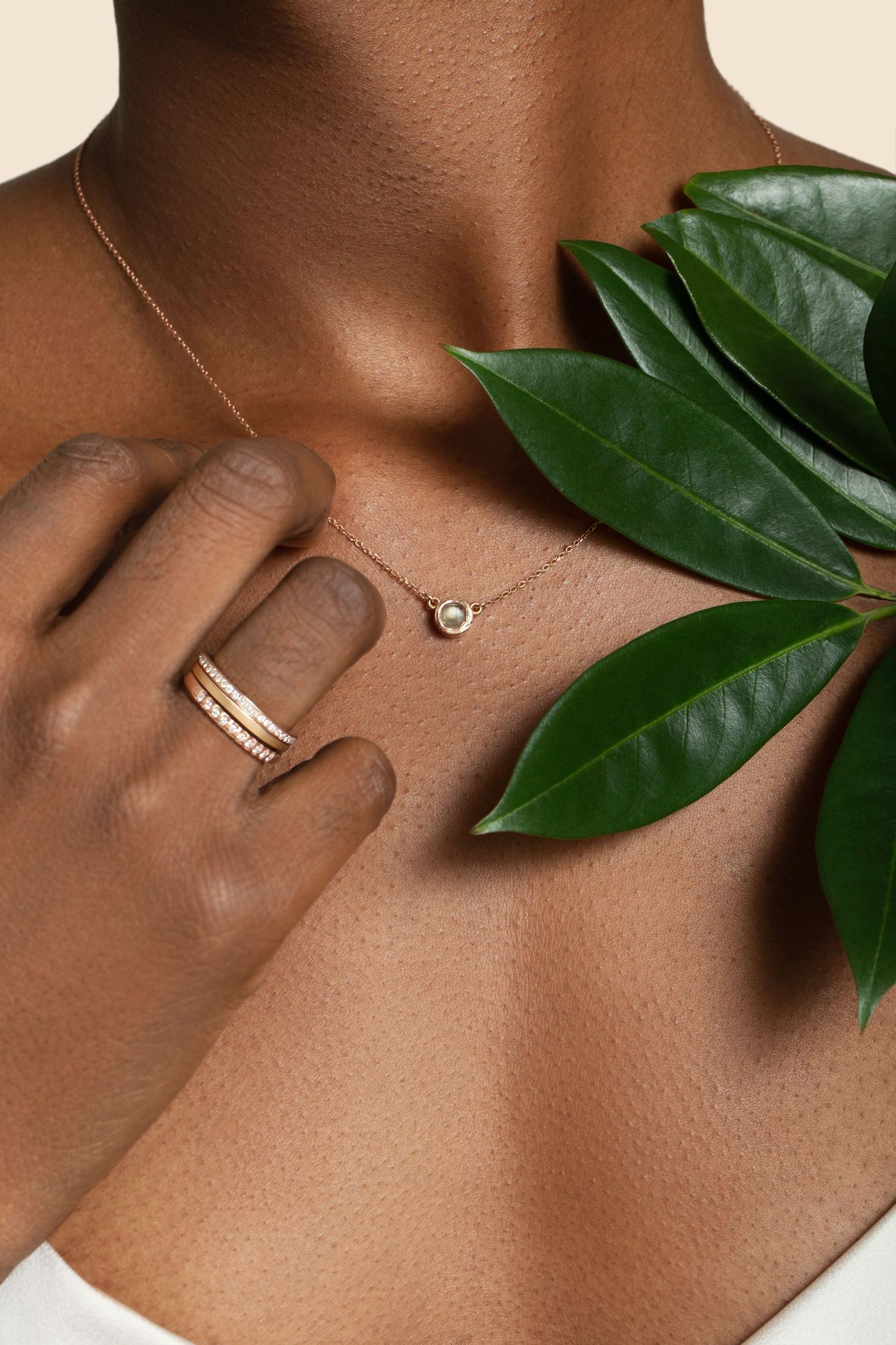 Close-up of a model wearing a delicate gold diamond necklace and stacked gold rings, posed with a green leaf.