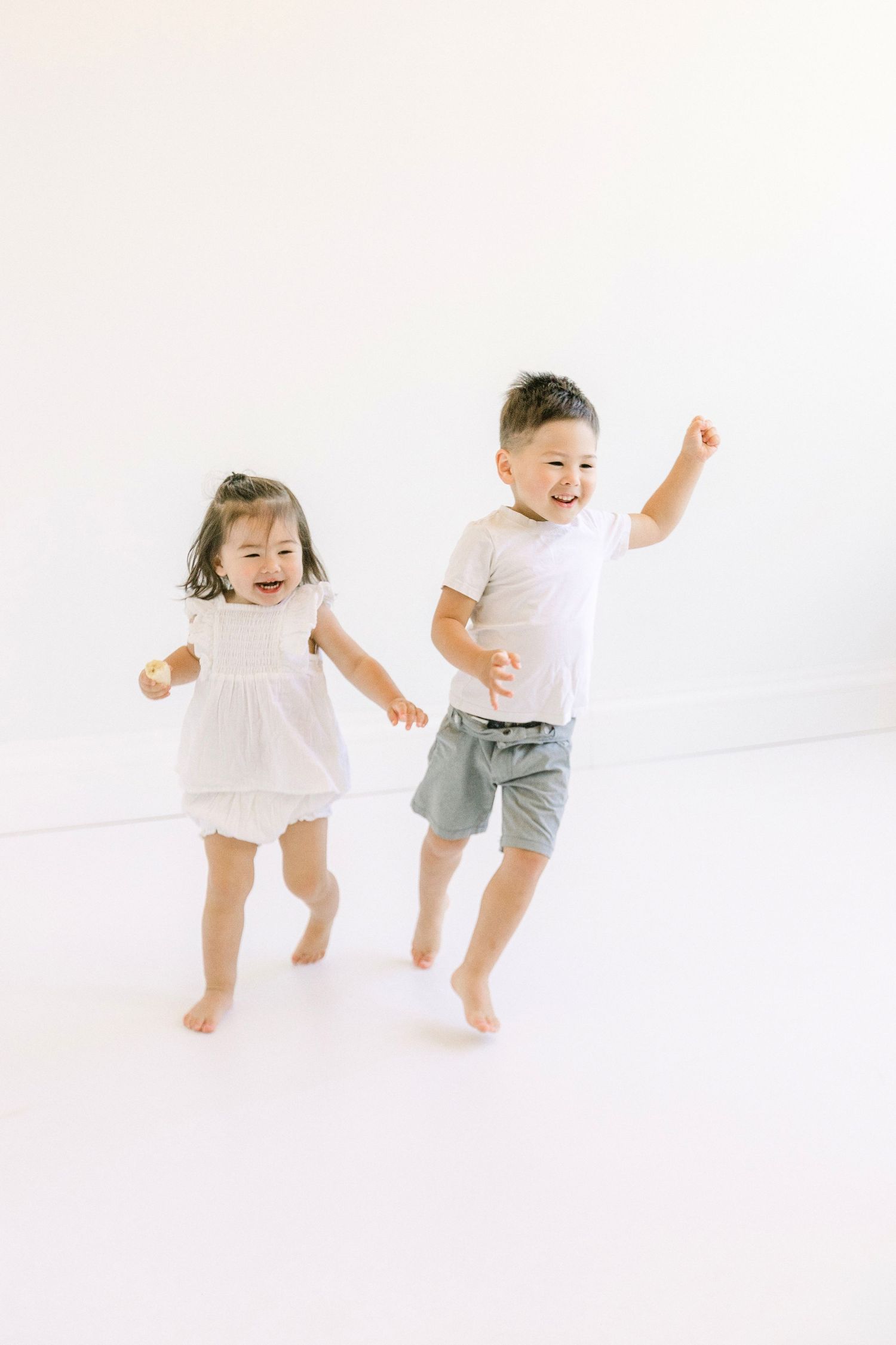 Two happy young Asian children jumping and laughing in a bright white studio, girl in white dress and boy in gray shorts.