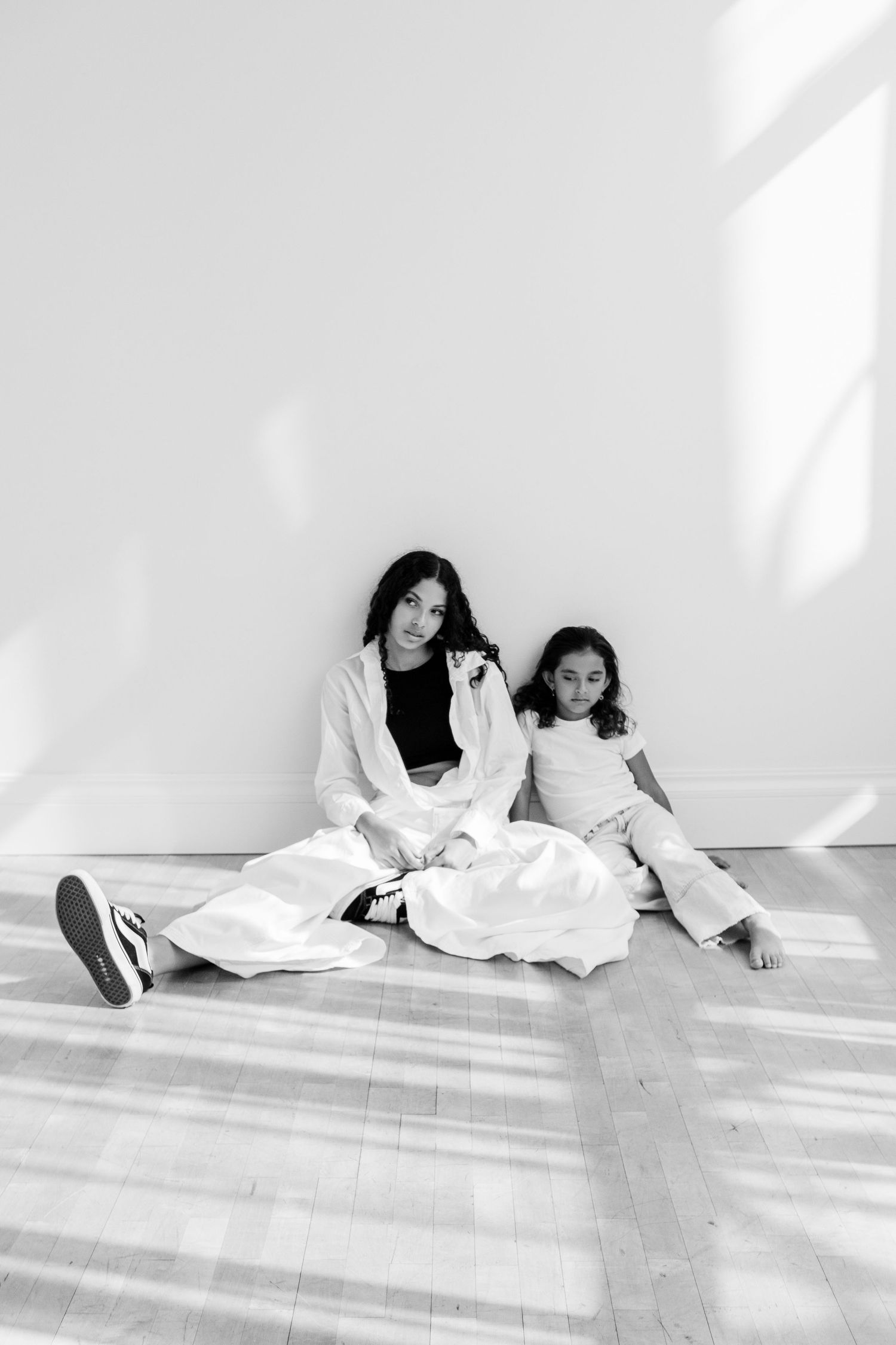 Two sisters sit together against a white wall in a black and white portrait photo.
