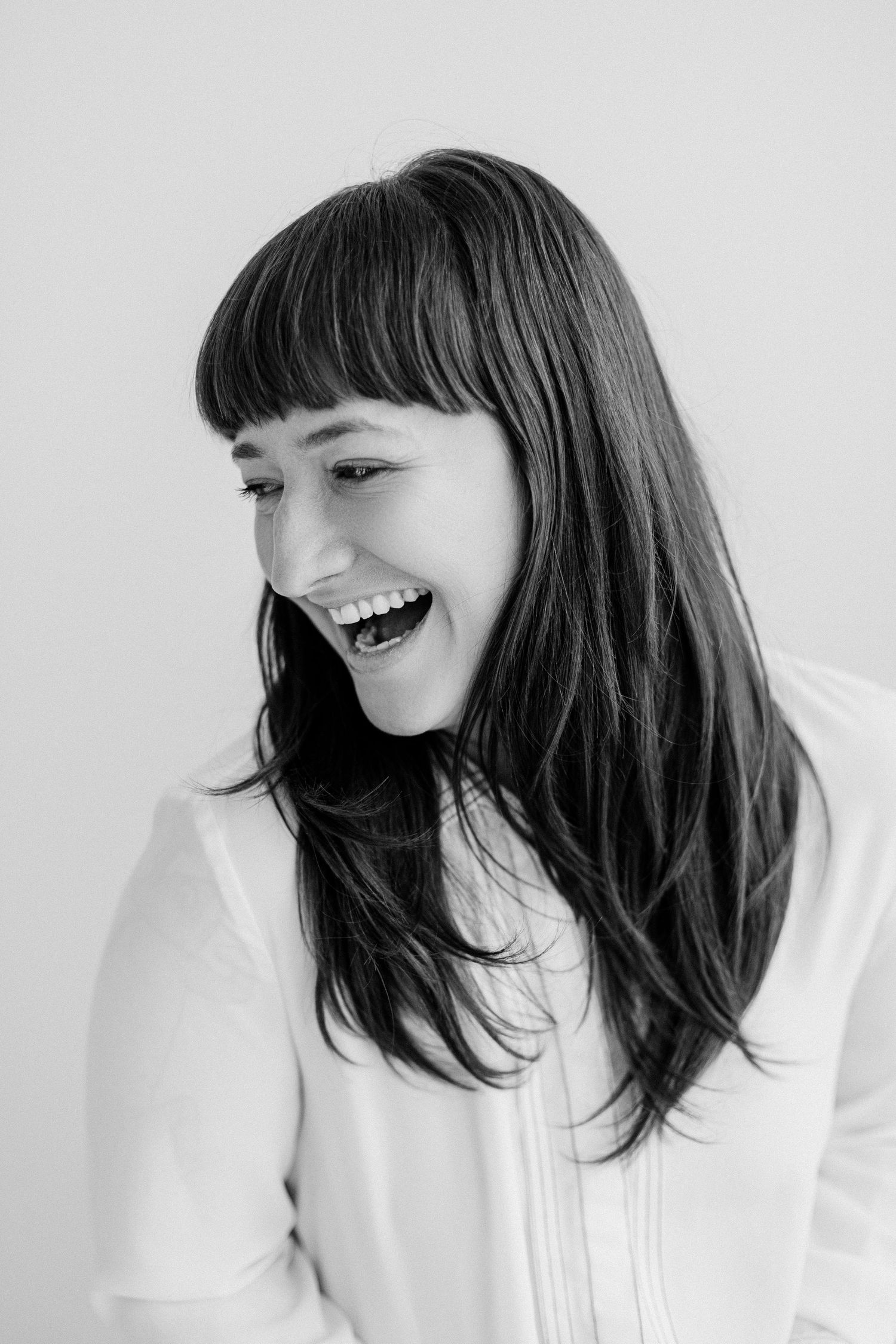 Black and white portrait of laughing brunette woman with bangs in white top against light background.