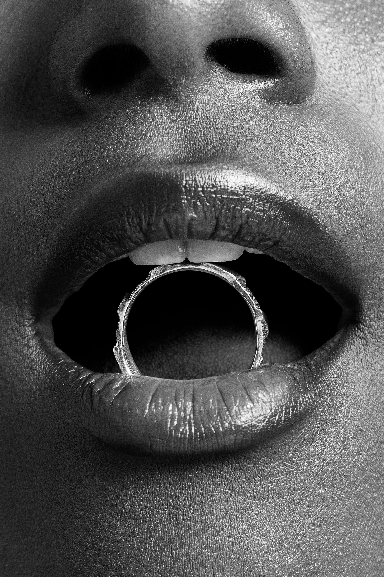 Black and white close-up of lips holding a silver ring in mouth, bold beauty editorial jewelry photography.