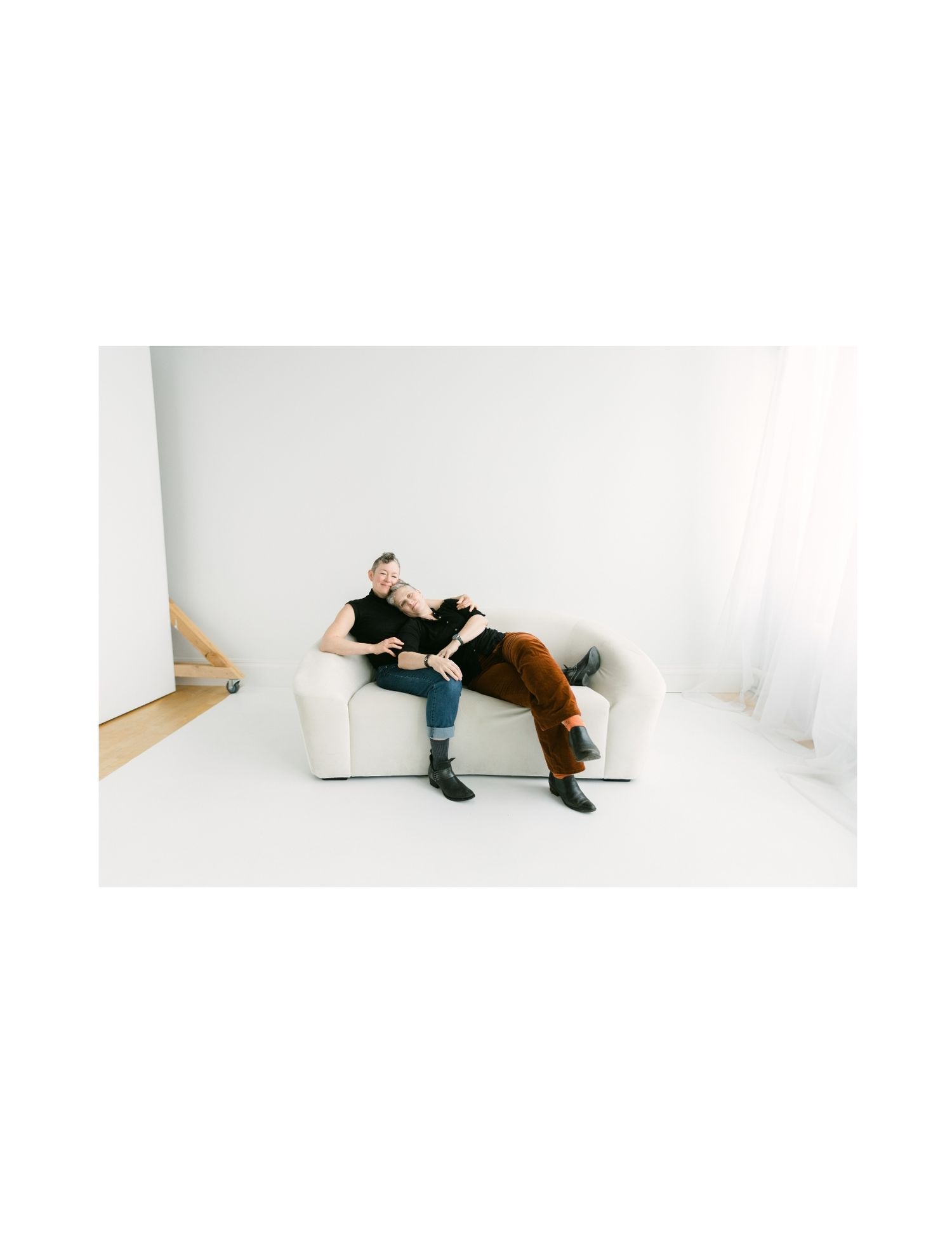 Two women relaxing together on a white couch, one resting her head on the others lap in a studio.