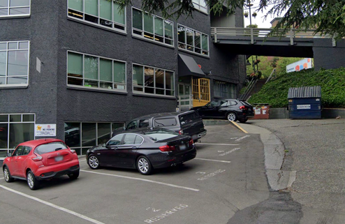 Gray commercial building with parking lot featuring multiple cars, surrounded by trees and a bridge in background.