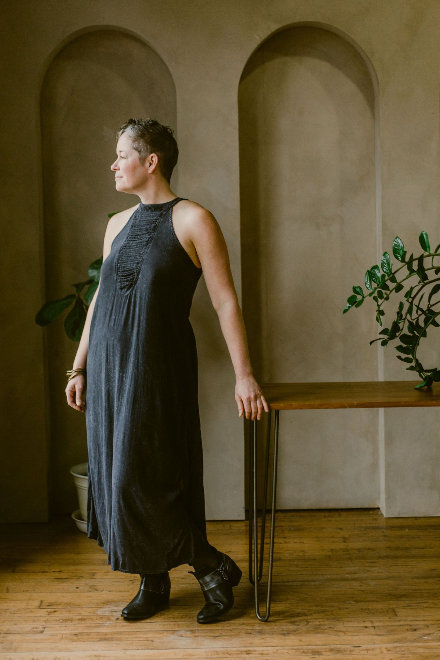 Woman in dark maxi dress and boots stands elegantly by a wooden table in a neutral-toned room with arched alcoves.
