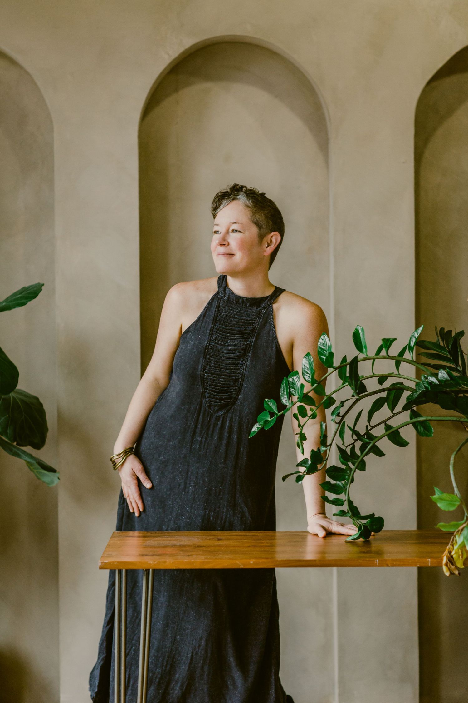 Woman in black halter dress stands by wooden table with plants, posed against arched stone wall backdrop.