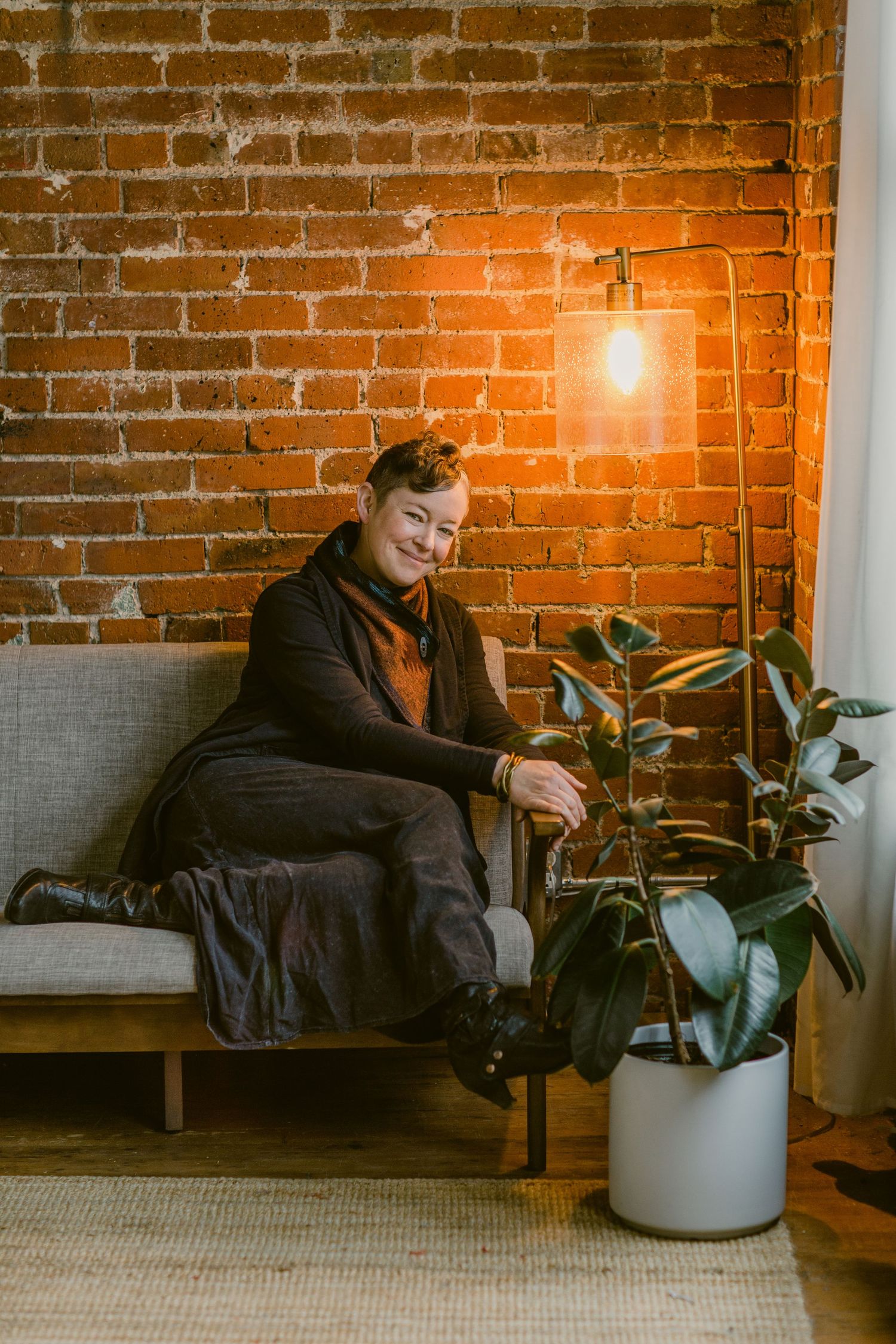 Person sitting on a couch near a brick wall, warm lamp light, and a rubber plant in a white pot.