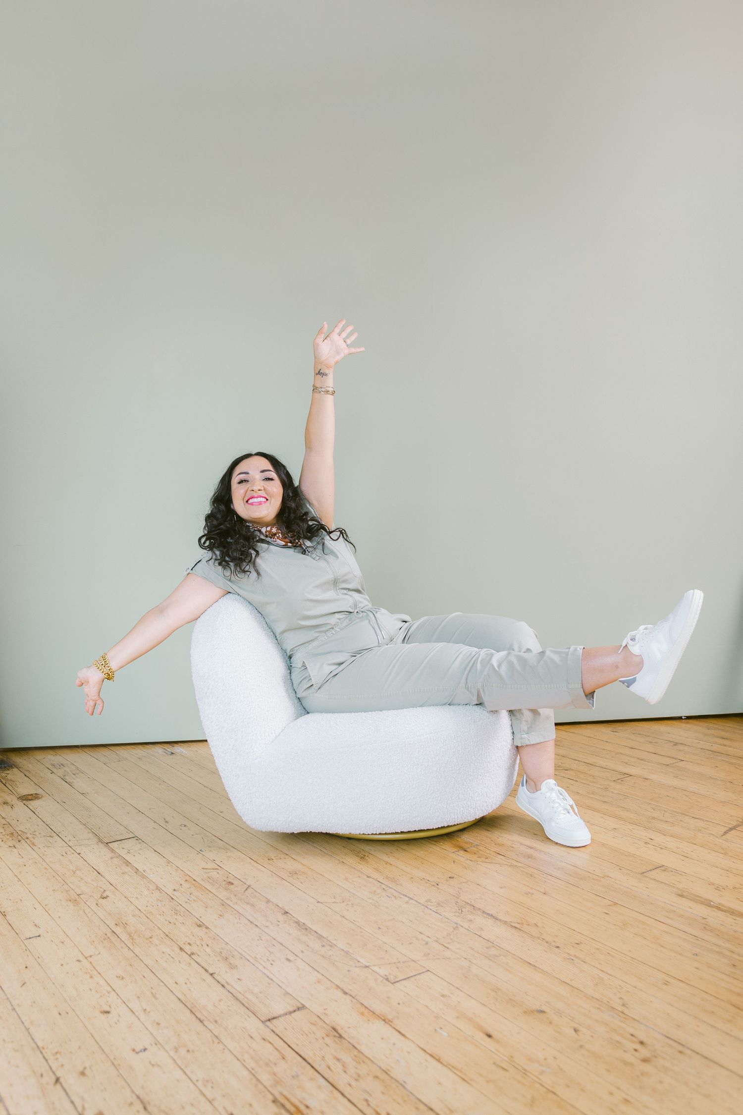 Woman in gray outfit sitting playfully in white modern chair with arm raised, on wooden floor against sage green wall.