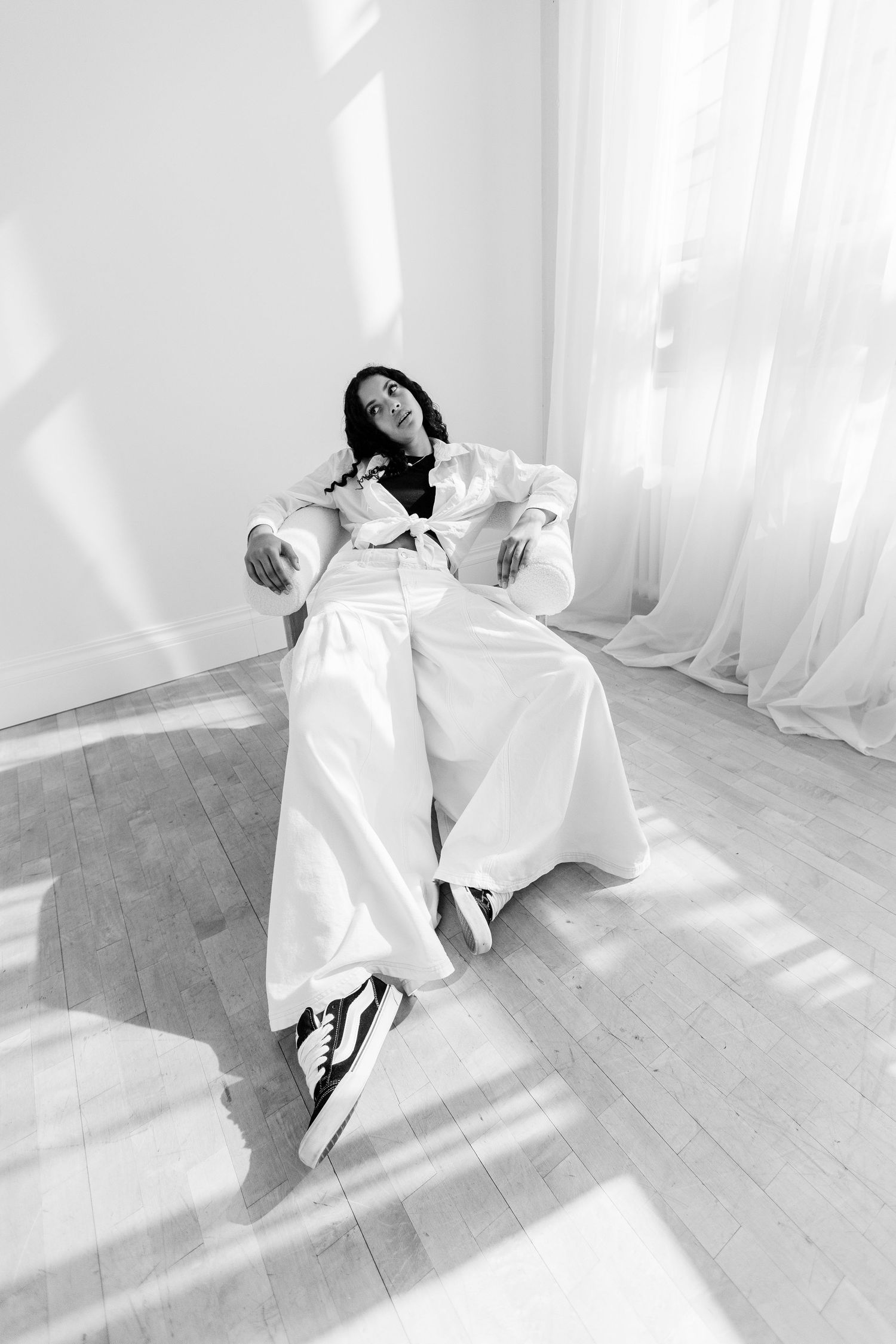 Young woman in white wide-leg pants and blazer relaxing in chair, black and white studio photo with natural light.