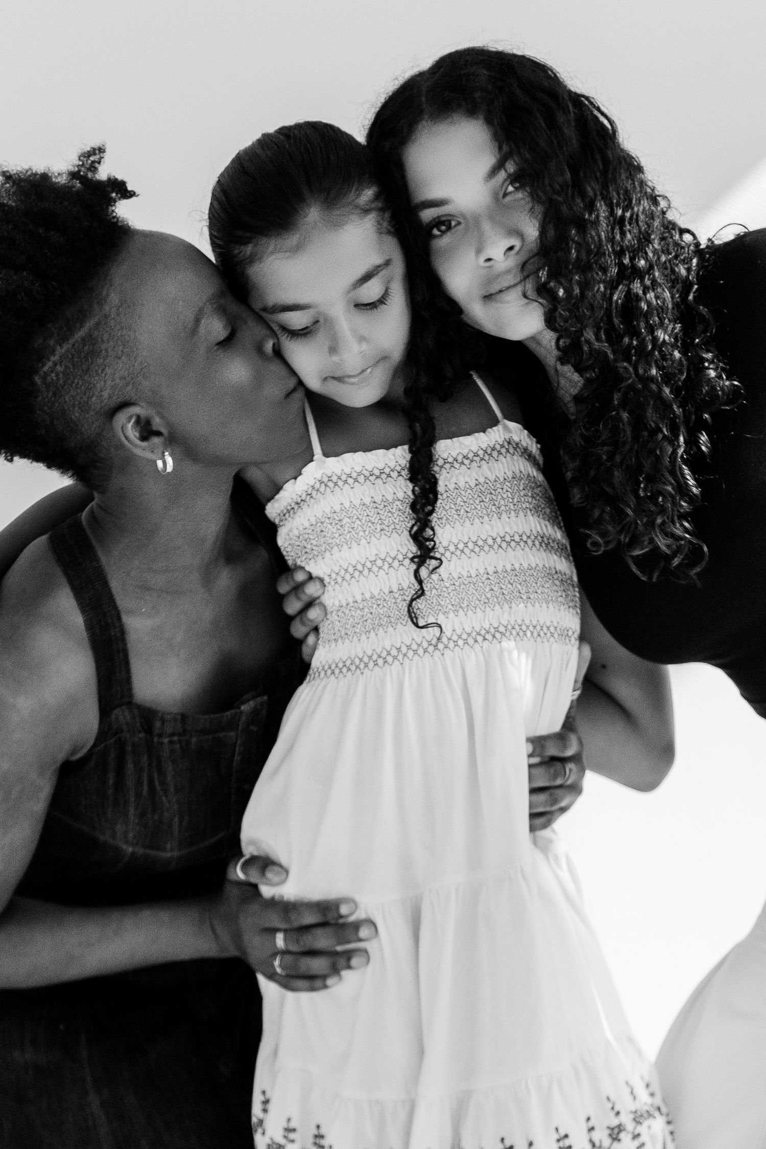 Black and white portrait of a mother kissing her daughter on the cheek while another daughter smiles nearby.