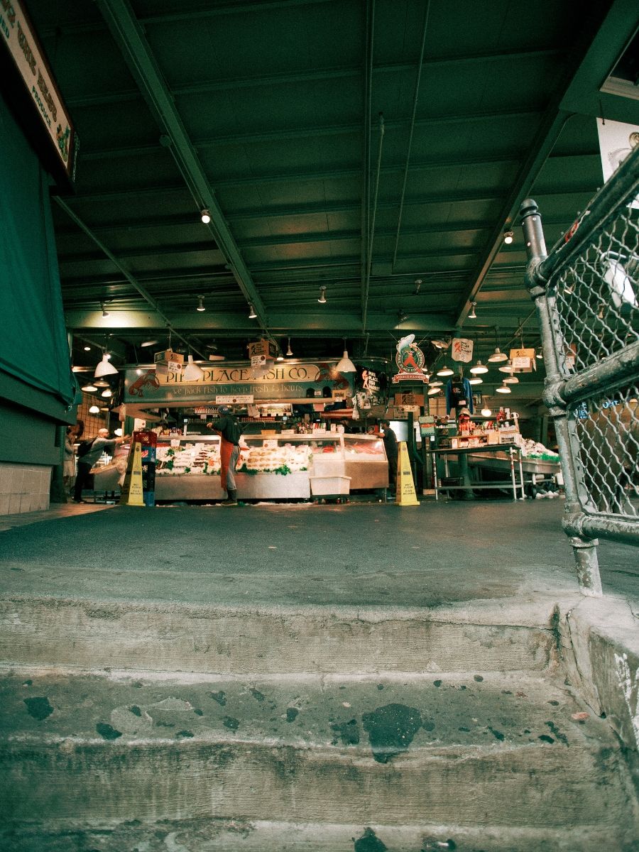 Pike Place Market fish stall with pink counter and hanging signs viewed from concrete steps in Seattle.